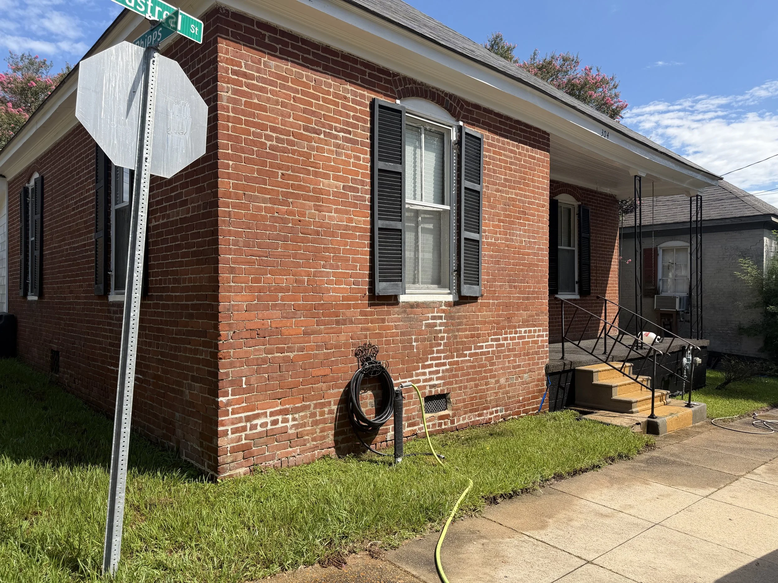 A red brick house with black shutters on the windows, a staircase with a black railing, and a yard with green grass. A street sign indicating 'Eustis St' and 'Quips St' is visible, along with some pink flowering trees in the background.