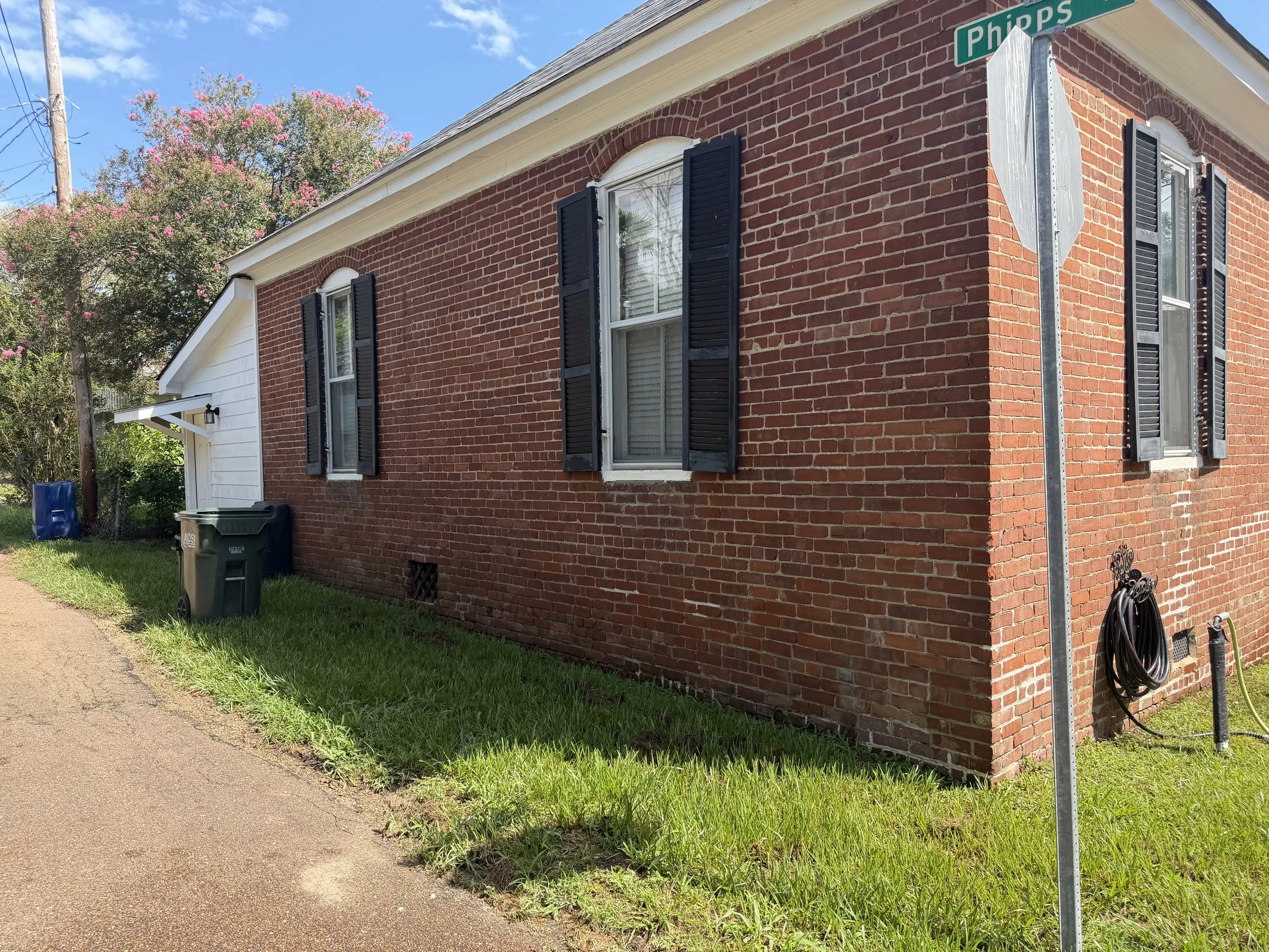 A red brick house with three windows with black shutters, white trim, and a front door with a small overhang. A street sign indicating 'Phipps,' a utility pole with wires, a tree with pink flowers, and some garbage bins are visible near the house.