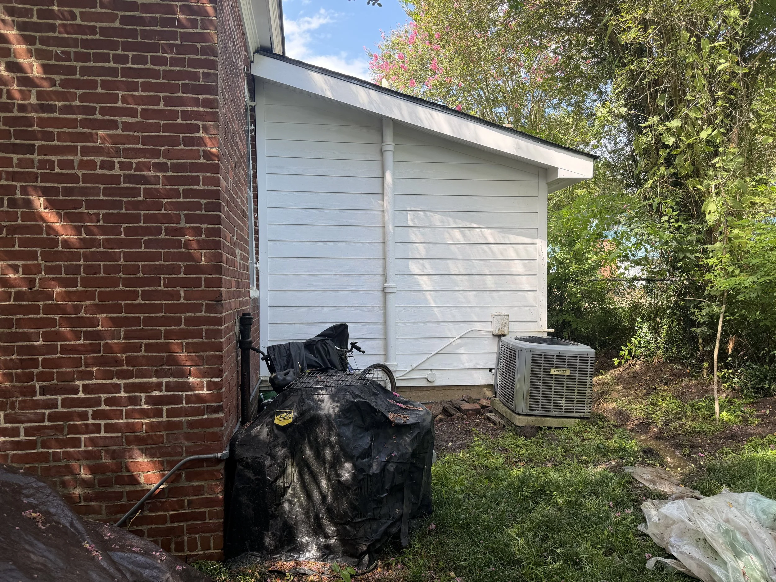Side view of a house showing part of a red brick wall, a white wall with siding, an outdoor air conditioning unit, a bicycle, large black trash bags, and surrounding green trees and grass.