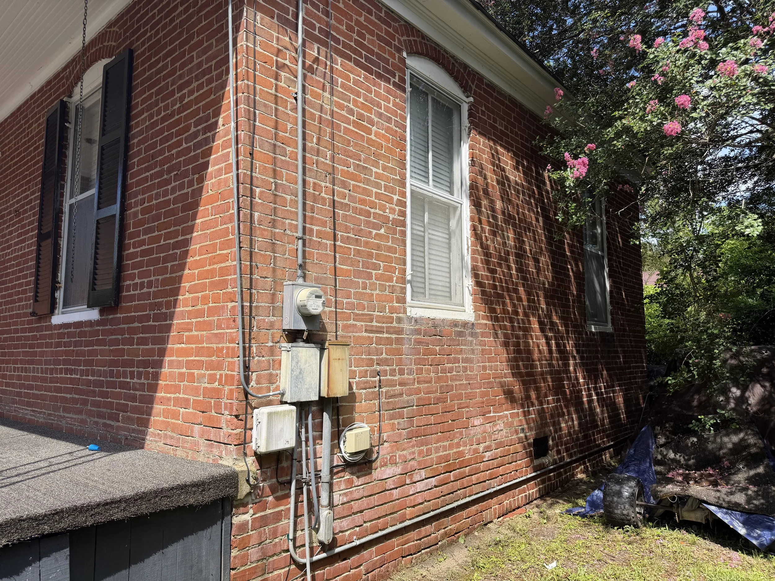 A brick house exterior with black shutters, white window frames, and electrical meters and conduits on the wall. There are pink flowering trees and green foliage nearby, with shadows cast on the wall.