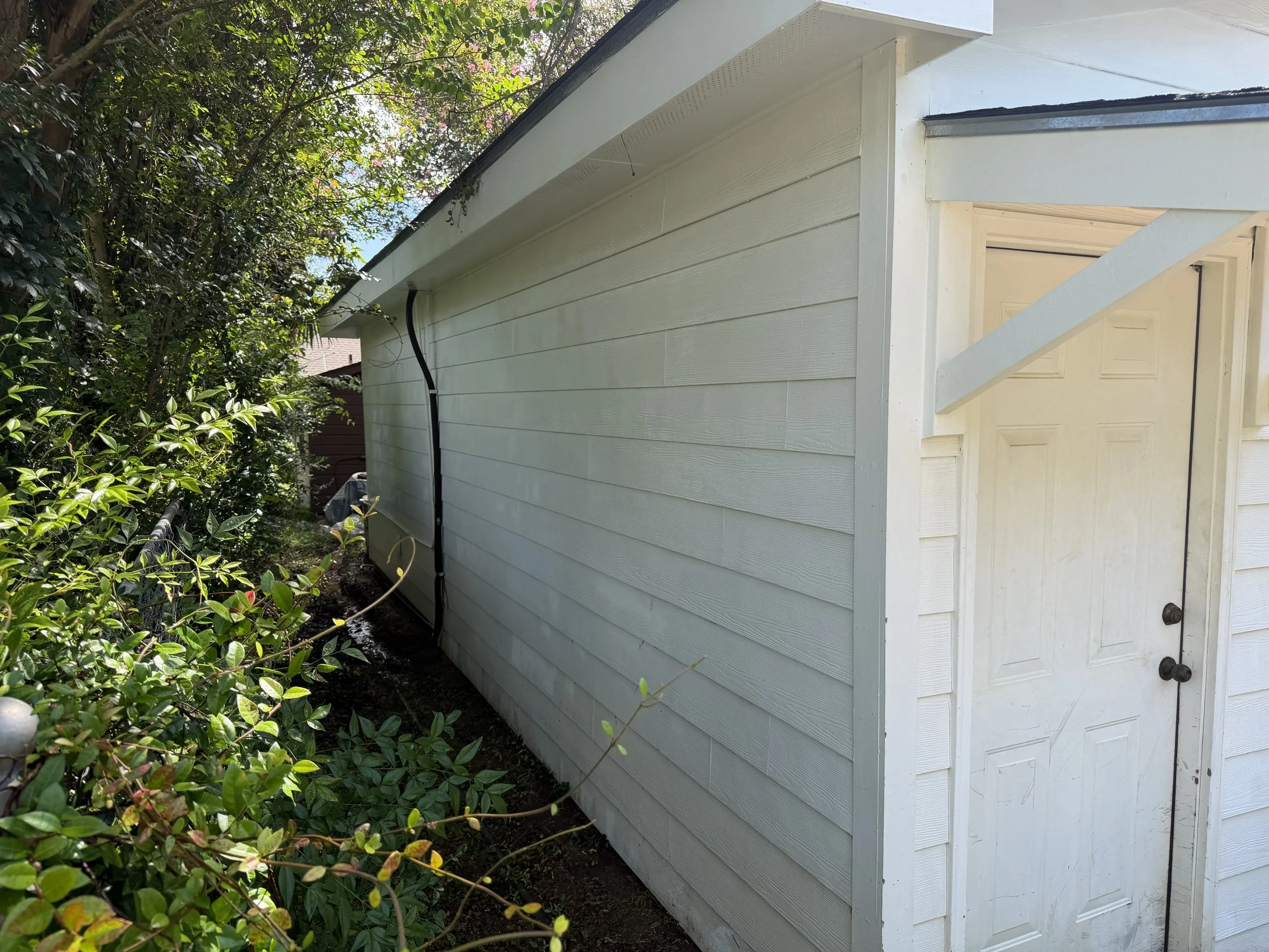 The side of a light-colored house with horizontal siding, partially shown door, and a black cable running along the exterior wall. Surrounding greenery with bushes and trees.