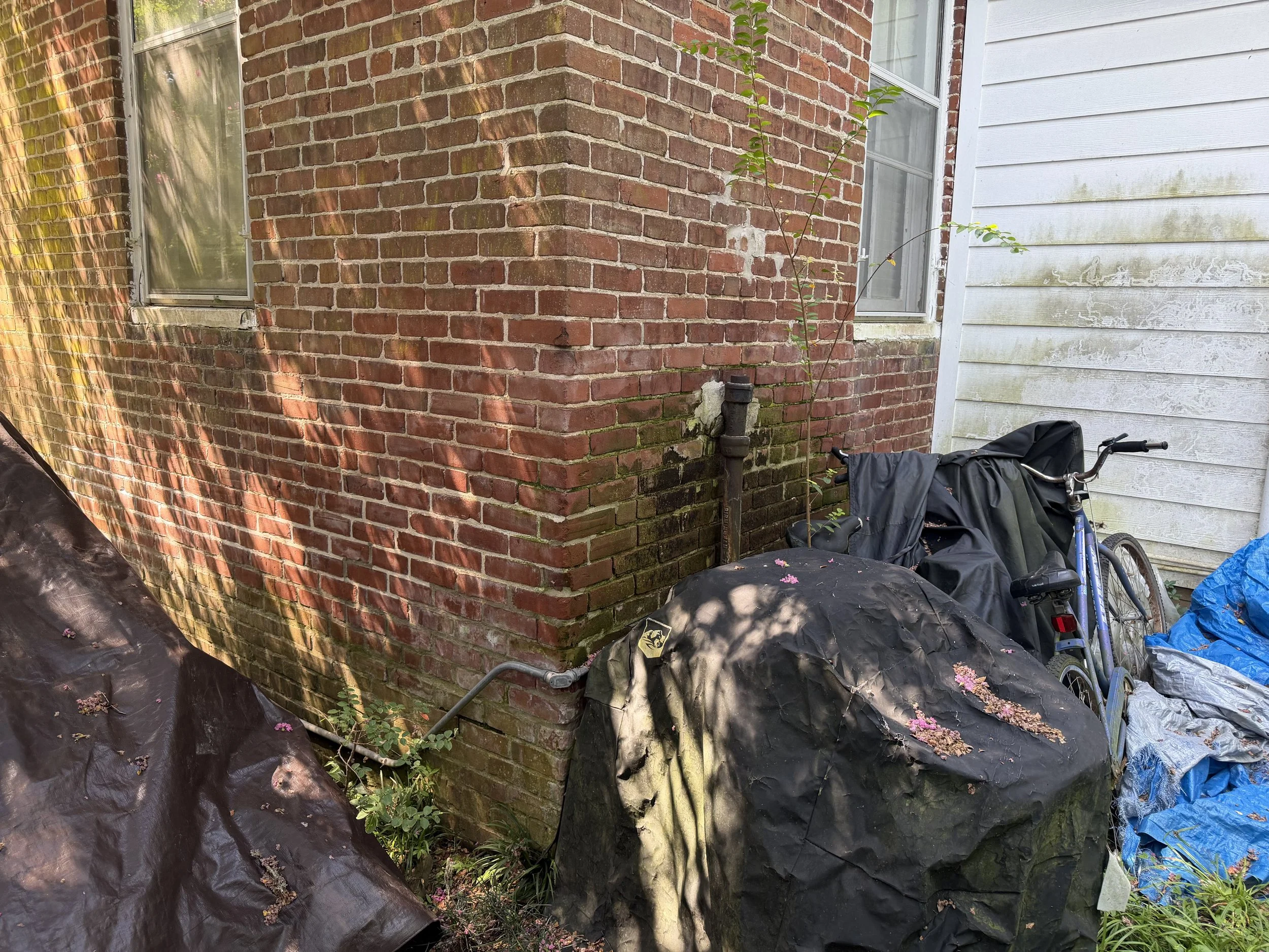 Brick corner and white siding with nearby plants and yard items before exterior cleaning.