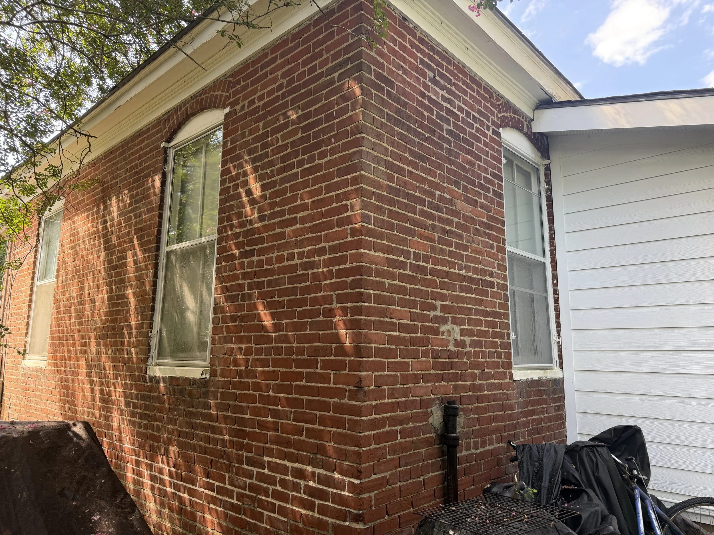Back corner of a brick house with windows, white siding, and a black drainpipe. There are trees with pink flowers and a blue sky in the background. There are a black grill cover, a black bag, and a bicycle in the lower right corner.