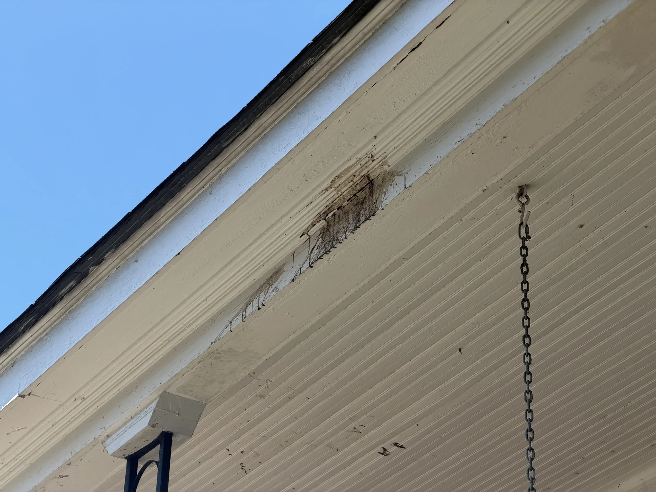 Close-up of the underside of a house porch or overhang with beige wood siding, a chain hanging down, and some dirt or mold near the edge. The sky is visible in the background.