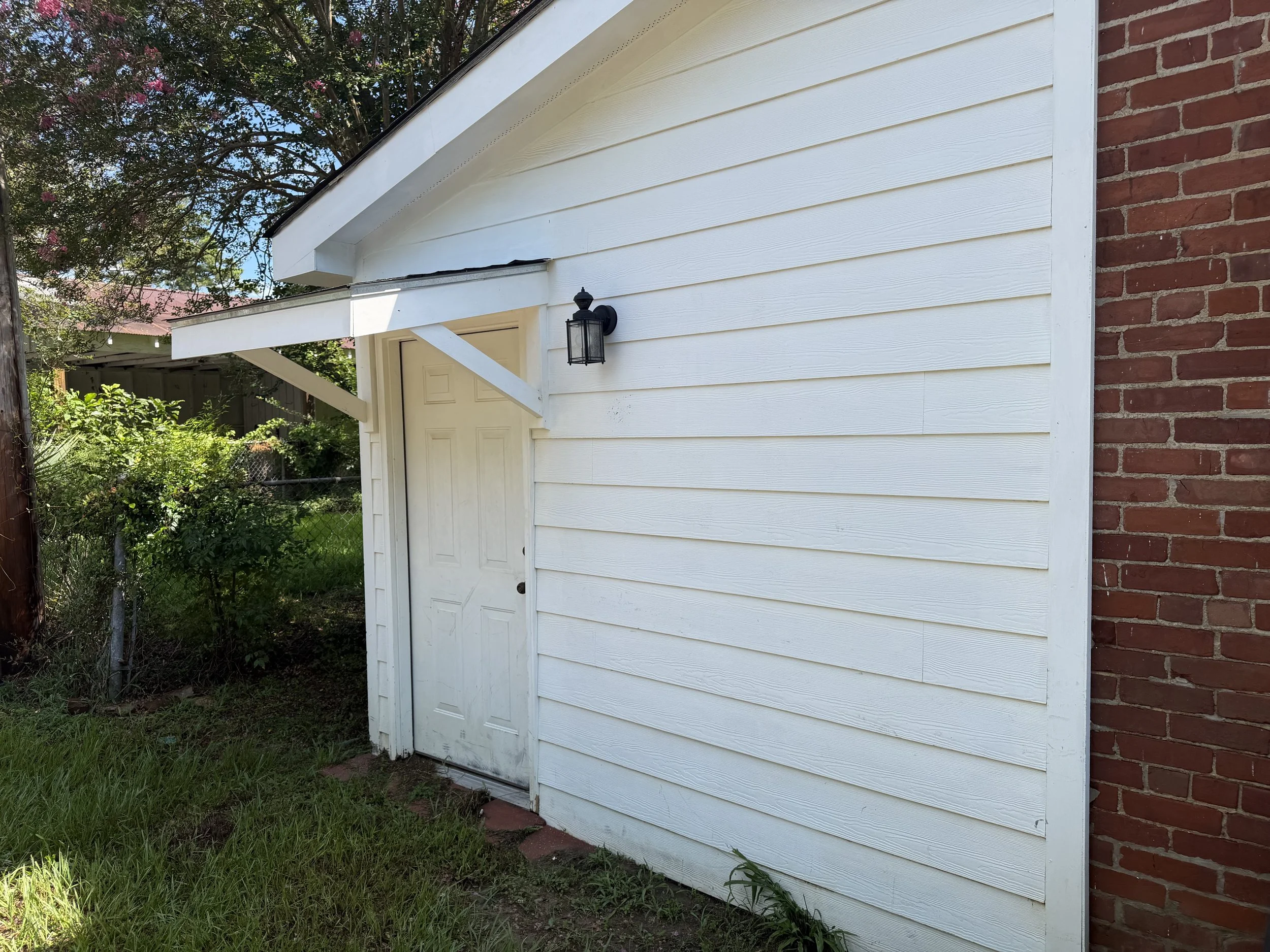Exterior view of a building with white siding and a brick wall, featuring a door and a black outdoor light fixture.