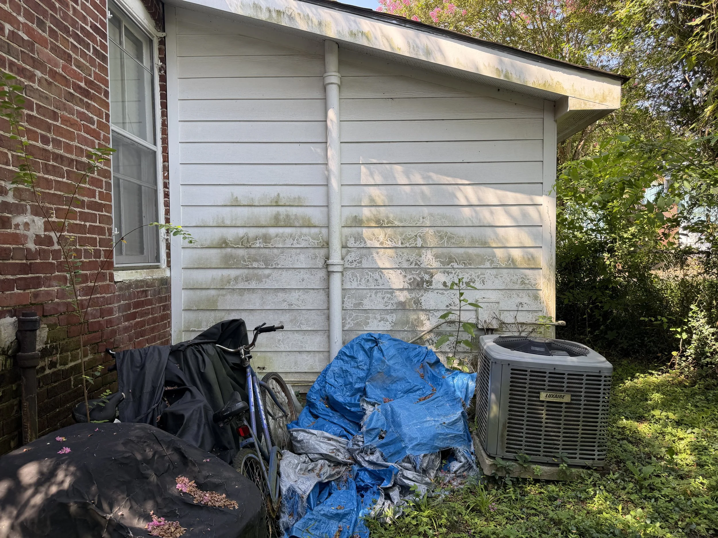 Backyard exterior wall with white siding, brick, AC unit, and items near the work area before cleaning.