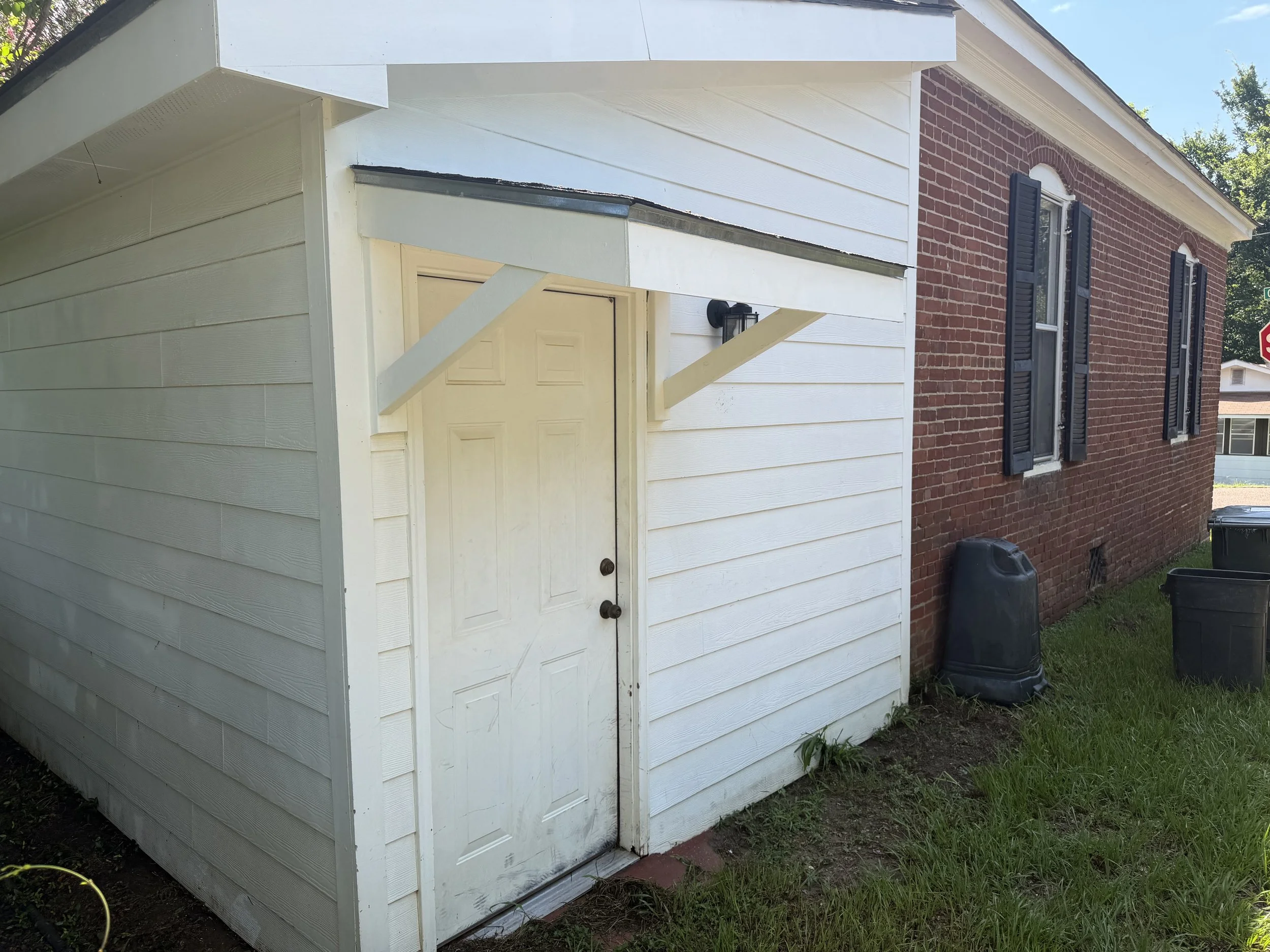 A small white shed with horizontal siding and a door, situated next to a brick house with black shutters, black outdoor light fixture, and window. There are black trash bins and a yellow garden hose in the yard.