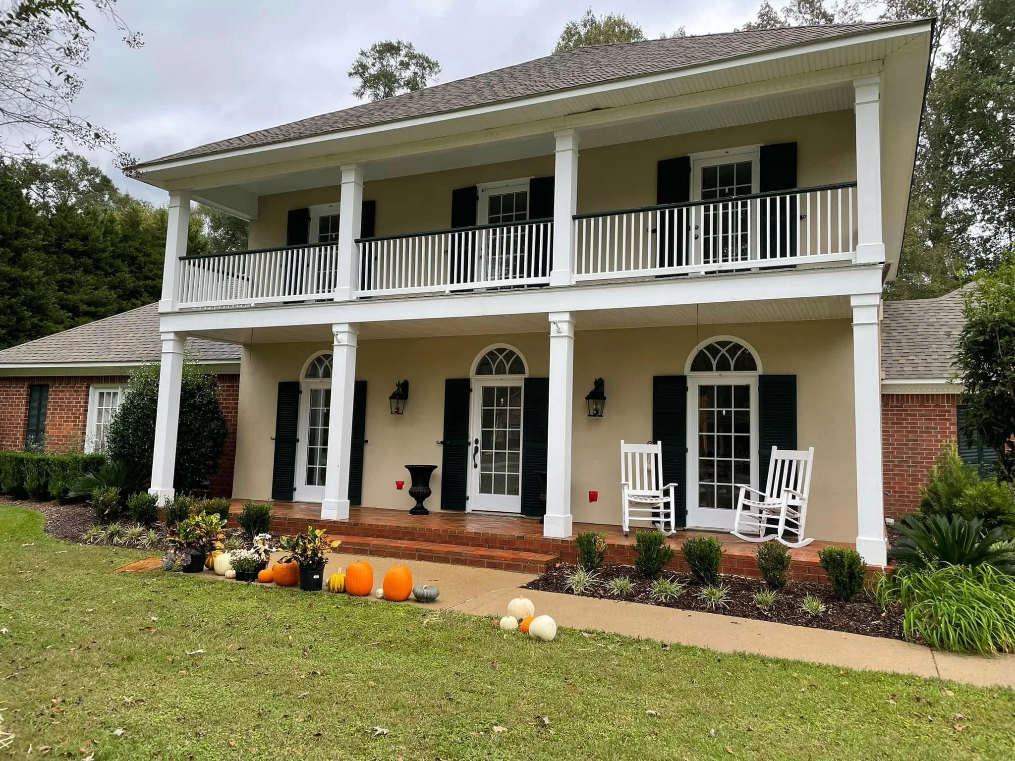 Front view of a two-story house with a large porch and balcony, decorated with pumpkins and plants outside, under a cloudy sky.