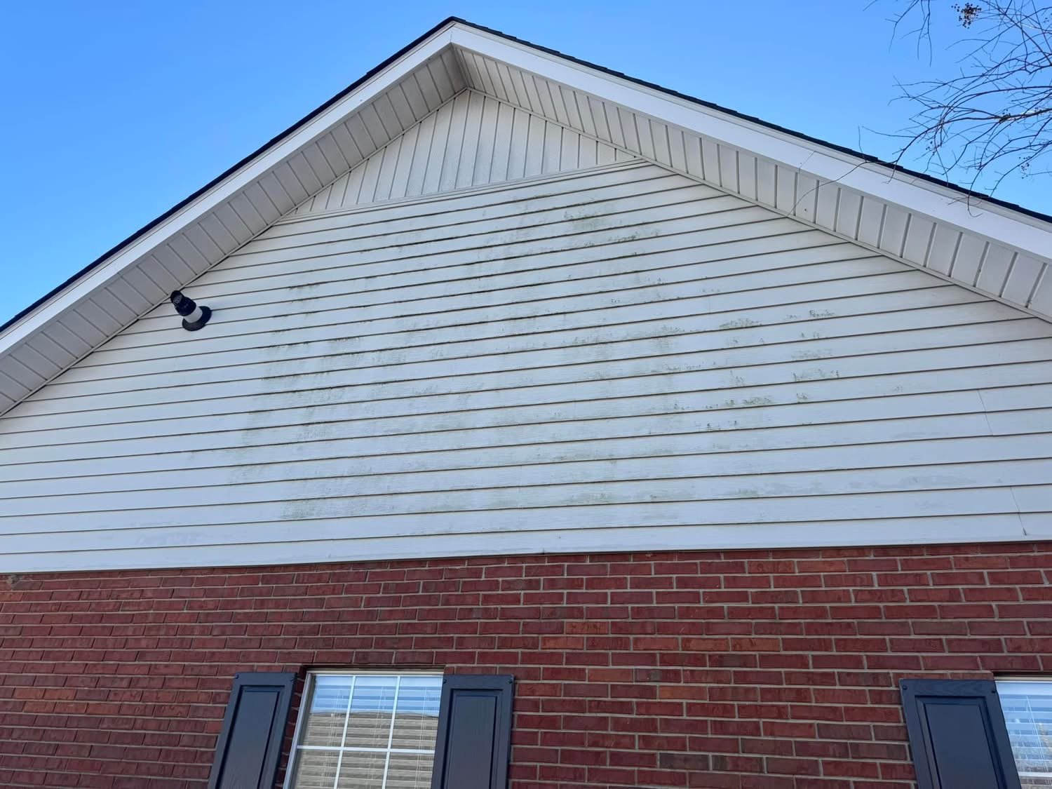 Close-up of a house's exterior showing part of the roof, white siding with dirt and grime, a black outdoor light, and a window with black shutters below.