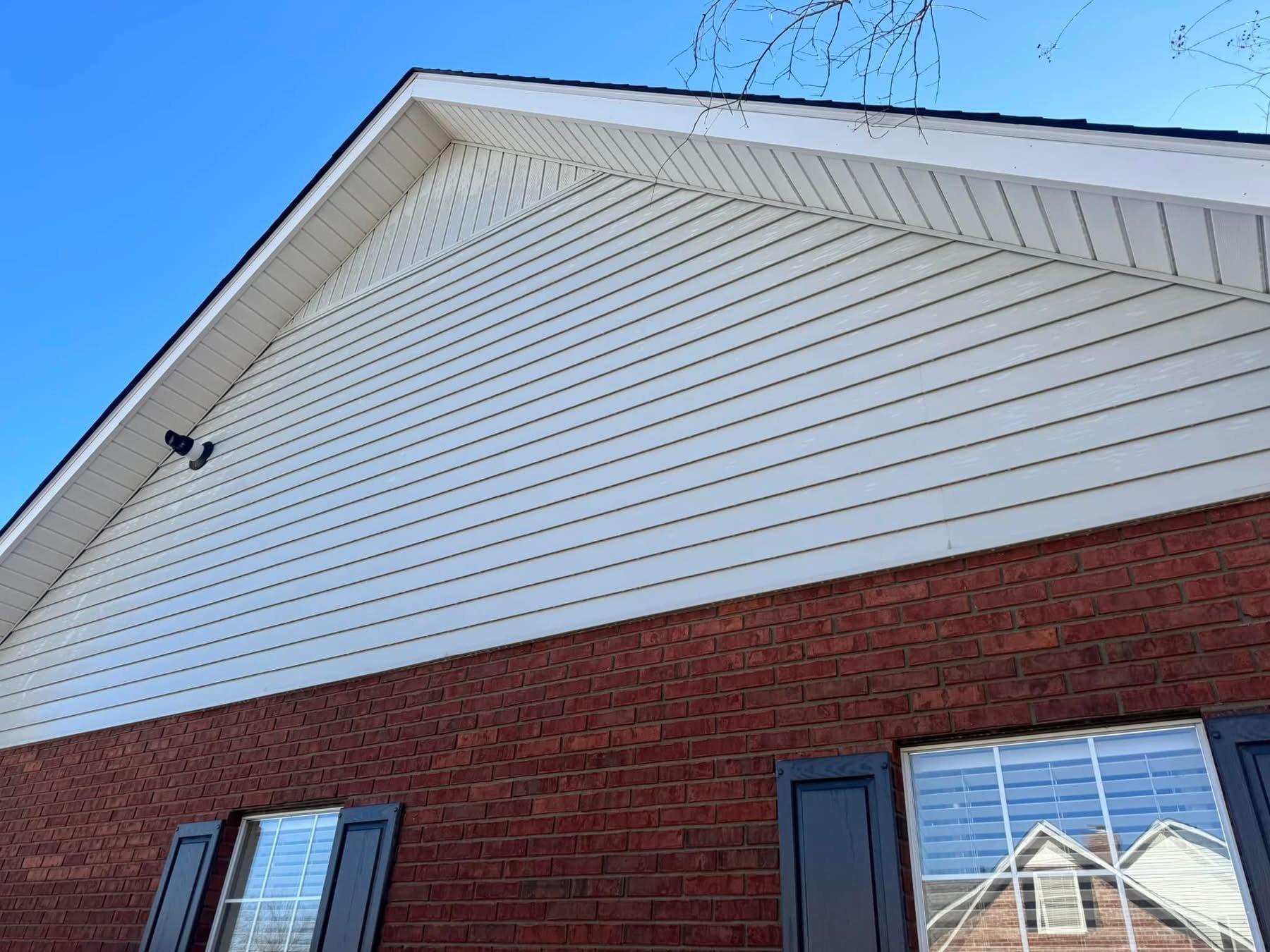 Close-up of the upper section of a house wall, featuring white horizontal siding on the upper half and red brick on the lower half, with windows having blue shutters and a security camera mounted on the siding.