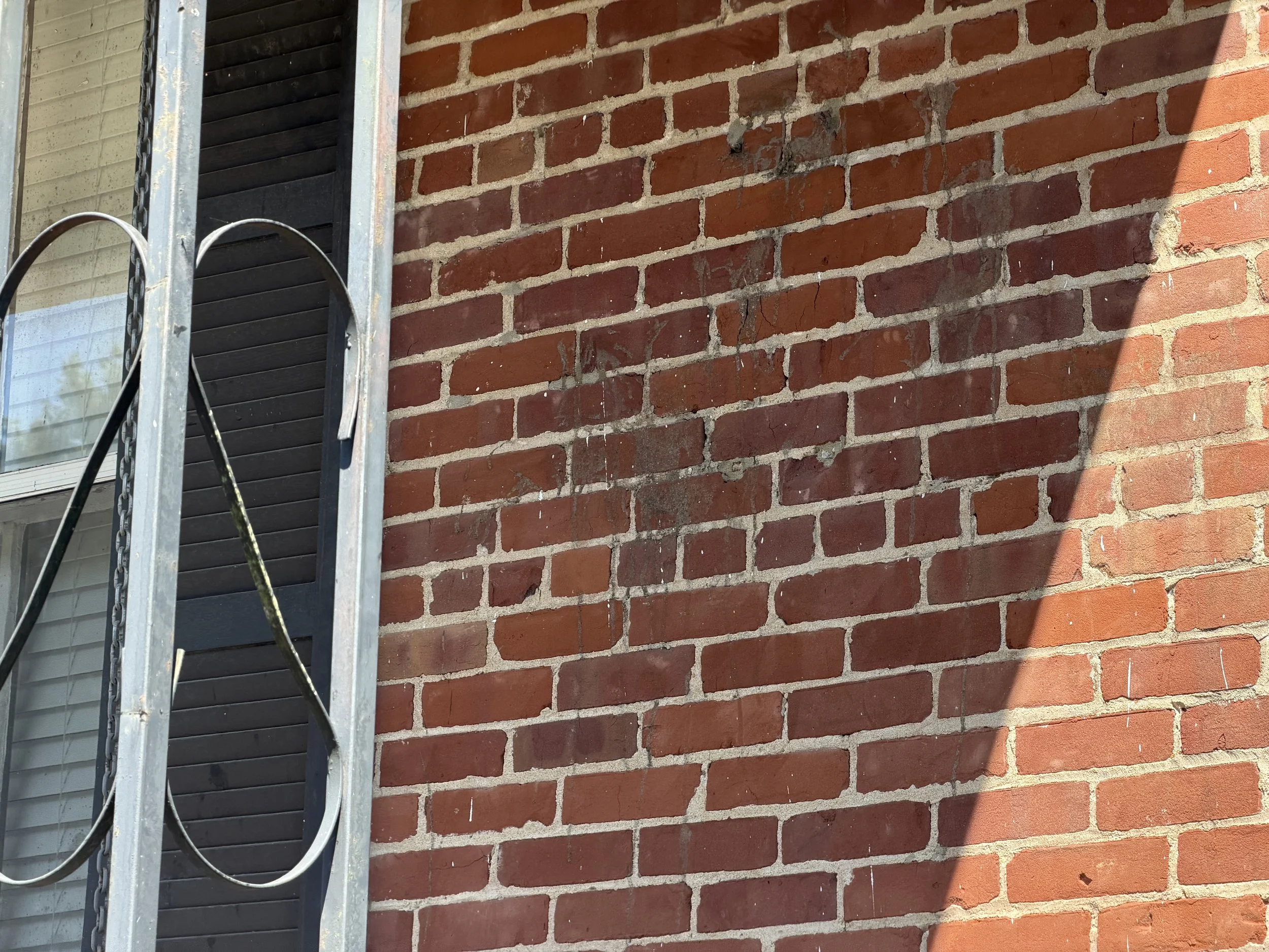 A brick wall with reddish-brown bricks and light gray mortar. Part of a metal frame with chains and black panels is visible on the left side, with the panel slightly open revealing a window with blinds behind it. Sunlight and shadows are cast on the 