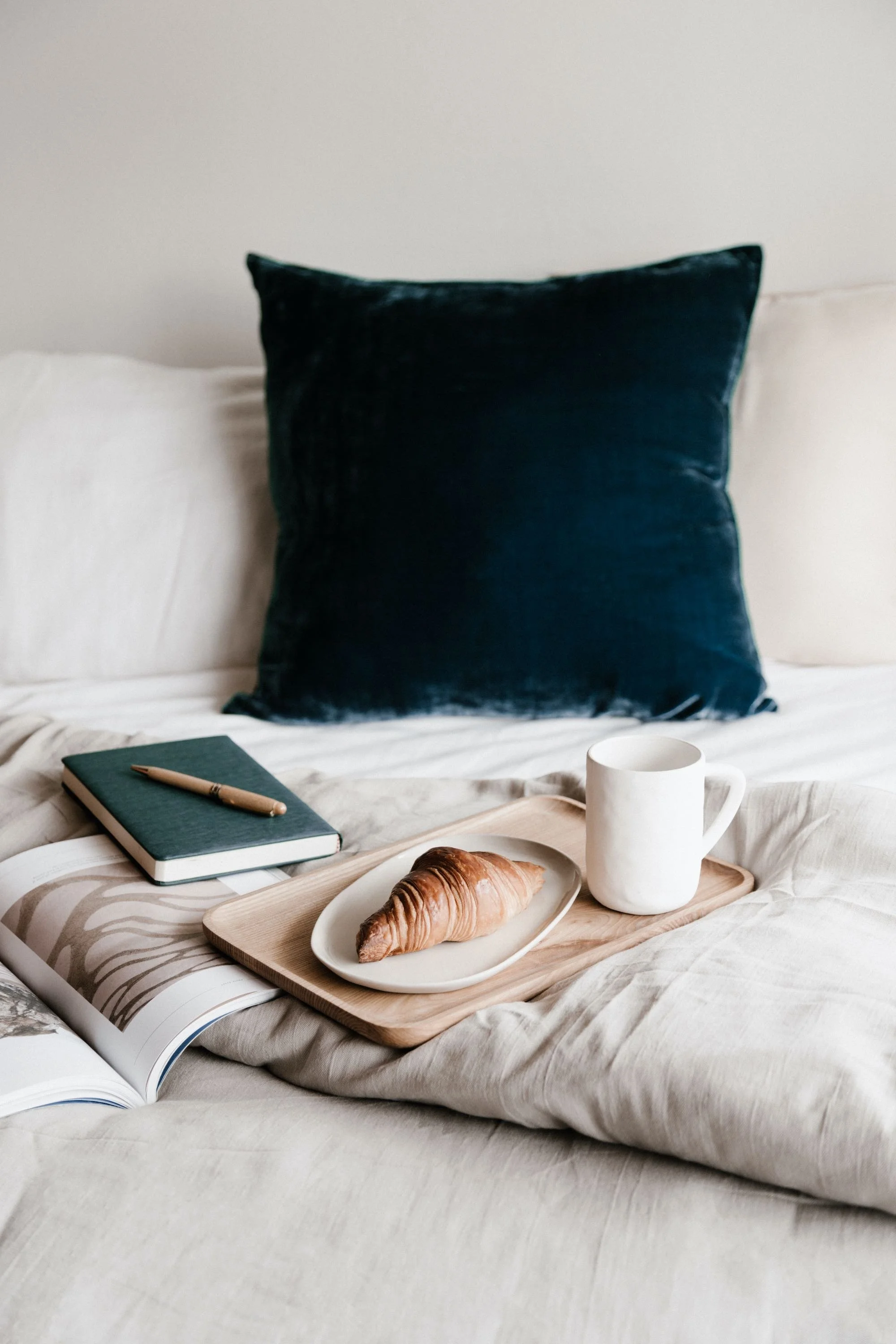 Croissant on a plate with a mug on a wooden tray, a notebook with a pen, and an open book on a bed with a dark blue pillow.