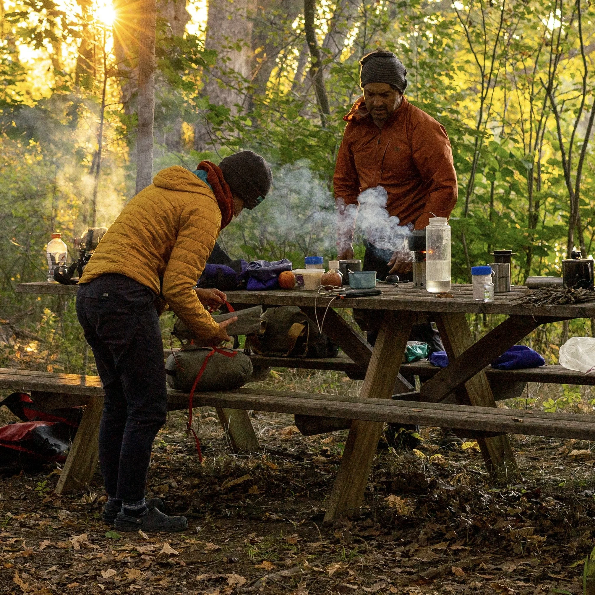 Two people cooking dinner at a campsite picnic table