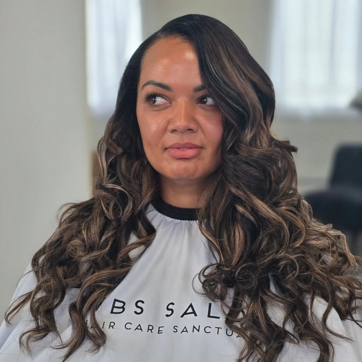 Woman with long, curly hair sitting in a salon chair, wearing a salon cape with text.
