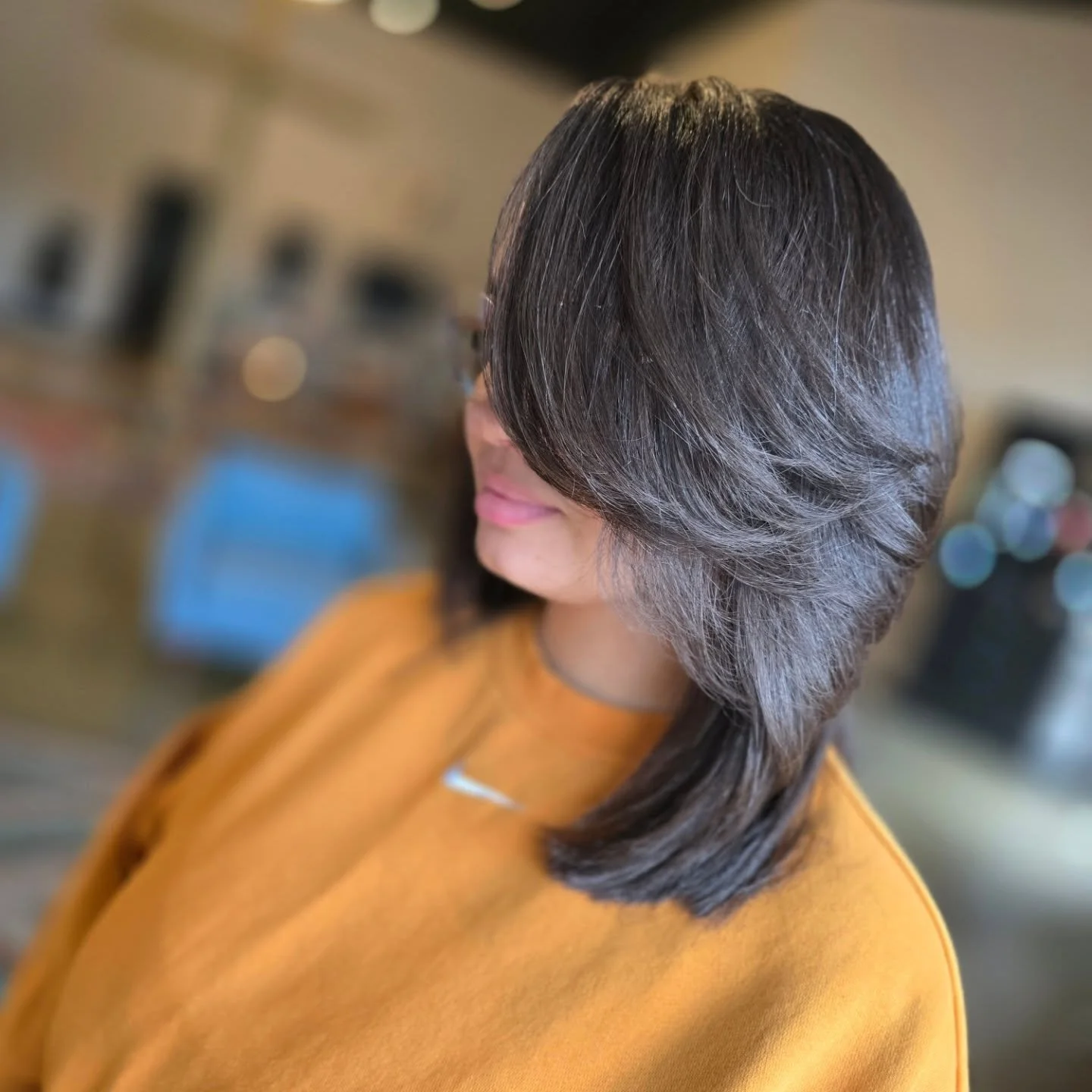 Profile of a woman with shoulder-length dark hair wearing an orange top, with a blurred background.