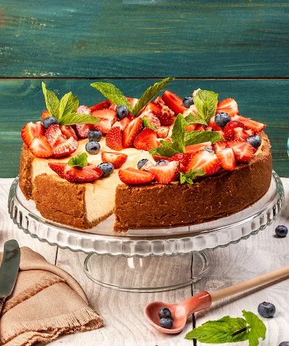 NY style cheesecake topped with sliced berries and mint leaves on a clear cake stand