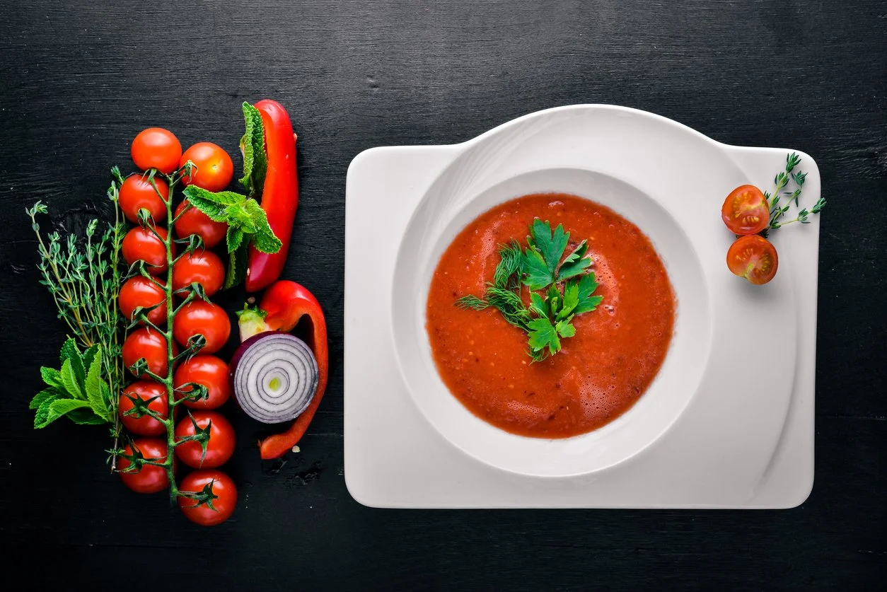 a white, geometric bowl filled with roasted red pepper and tomato soup, toped with parsley leaves, with cherry tomatoes on the vine, plus half a red onion and strips of red pepper, with sprigs of herbs