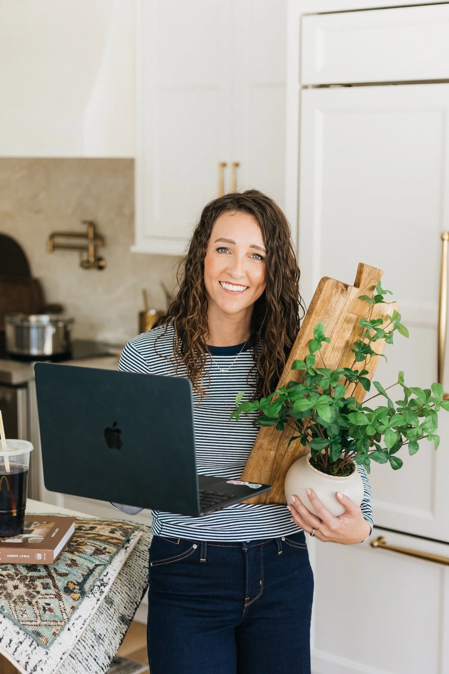 Chelsea Veurink Interior Design holding a potted plant in a white pot and a wooden cutting board, smiling in a kitchen with white cabinets and a table with a patterned rug, books, and drinks.