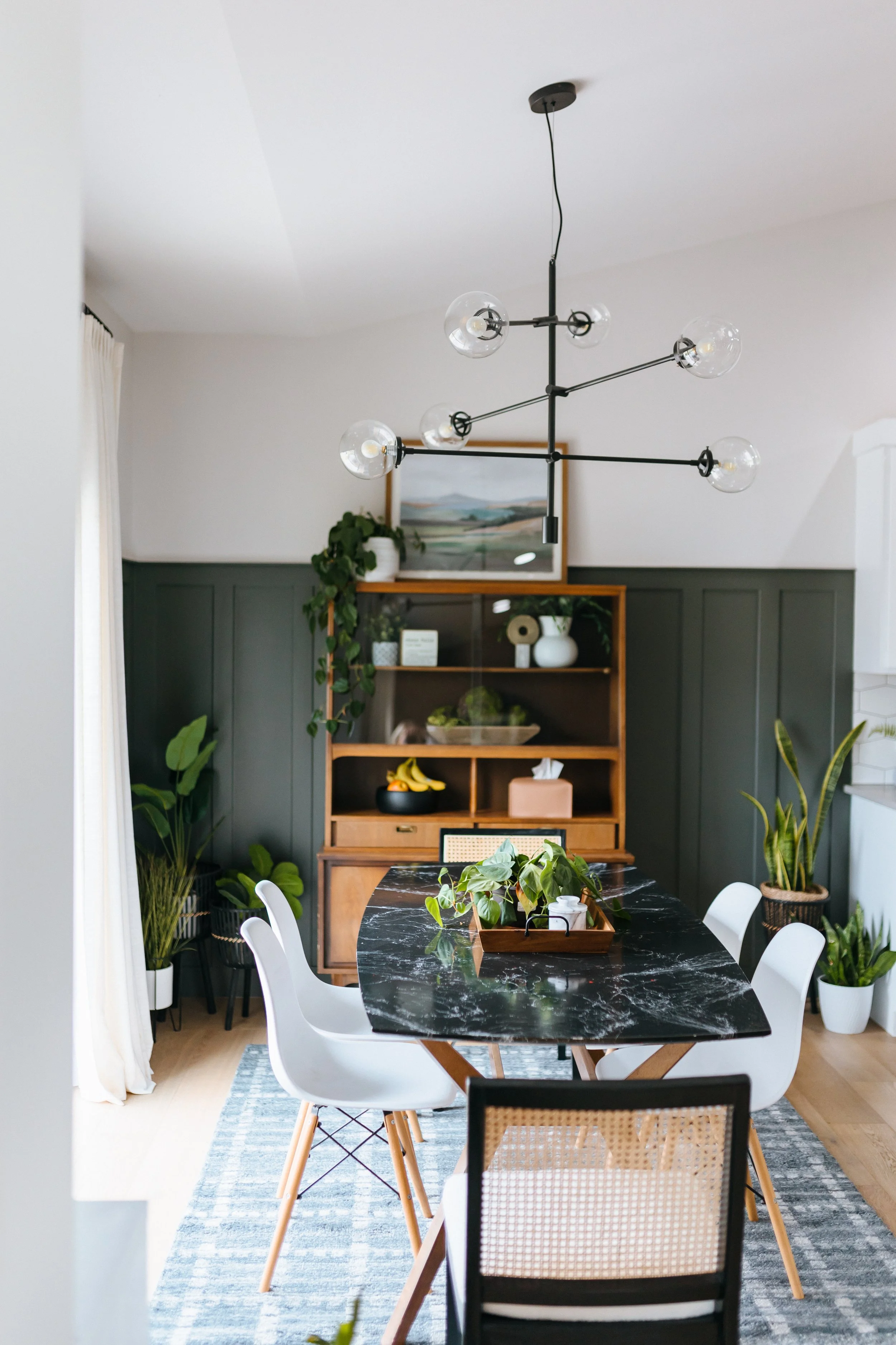 A dining area with a black and white marble table surrounded by white and rattan chairs. The room has a dark green accent wall behind a wooden bookshelf decorated with plants, artwork, and pottery. A floor-to-ceiling window with white curtains is on 