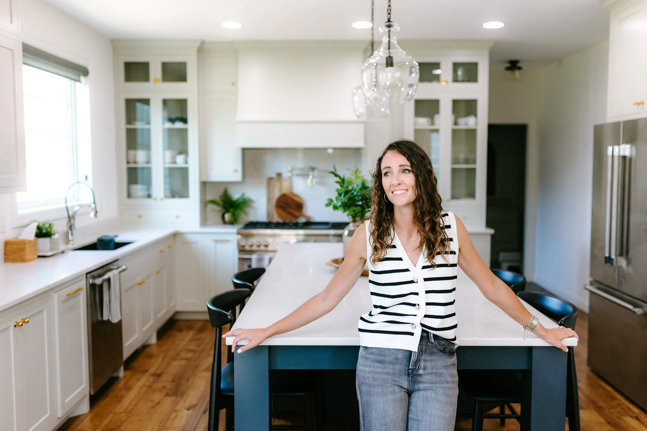 A woman with curly brown hair in a black and white striped sleeveless top and jeans leaning on a kitchen island with a smile, in a modern white kitchen with cabinets, a stove, and pendant lights.