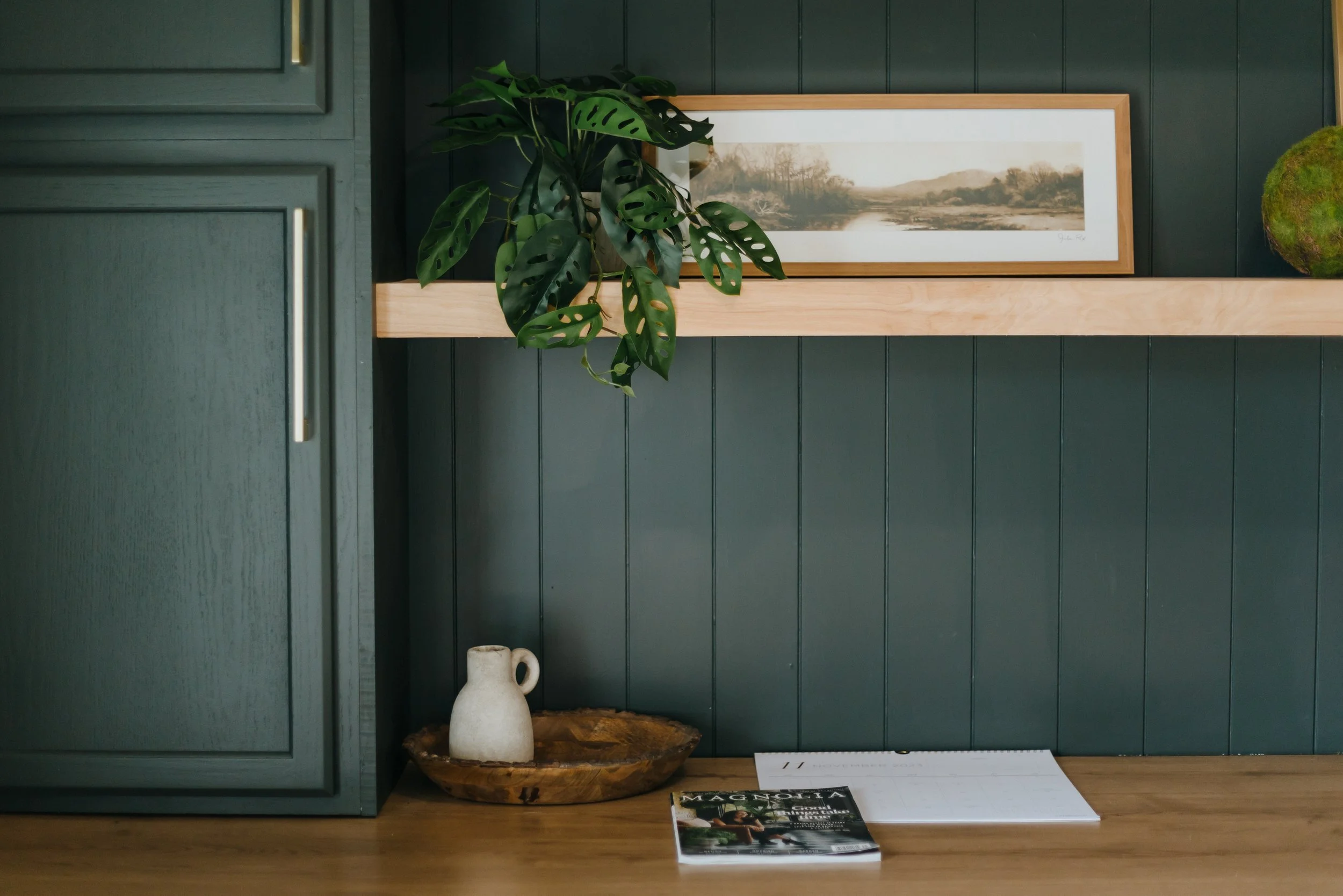 Interior scene with dark green-paneled wall, framed landscape artwork, a large green leafy plant on a wooden shelf, a white ceramic jug on a wooden table, and a magazine with a leaf on the cover.