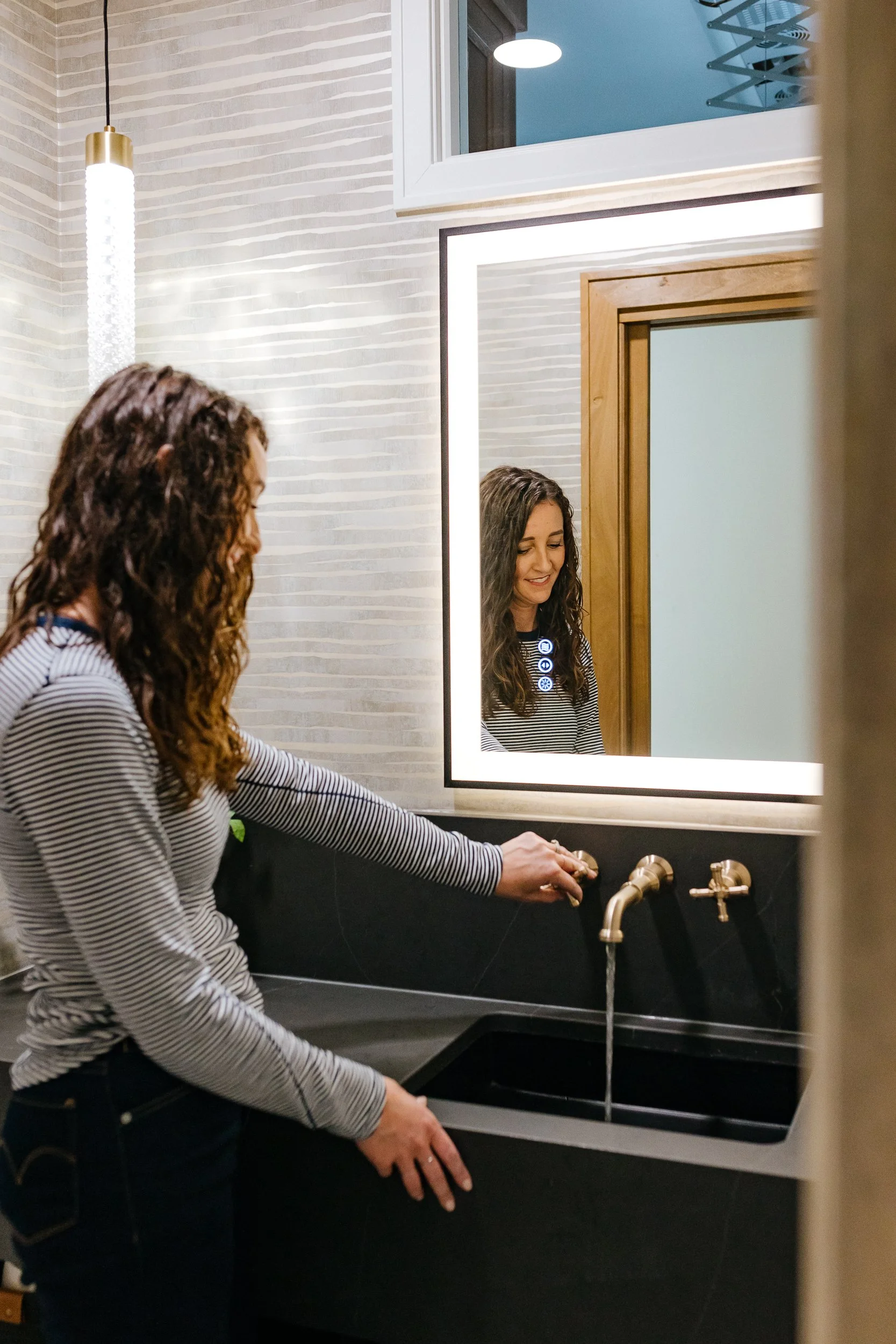 A woman with long curly hair wearing a striped shirt washing her hands at a black bathroom sink with gold fixtures. She is looking down and smiling at her reflection in a mirror with a bright white light frame. The bathroom has a textured white wall, a window with a view of an adjacent room or hallway, and gold and black elements.