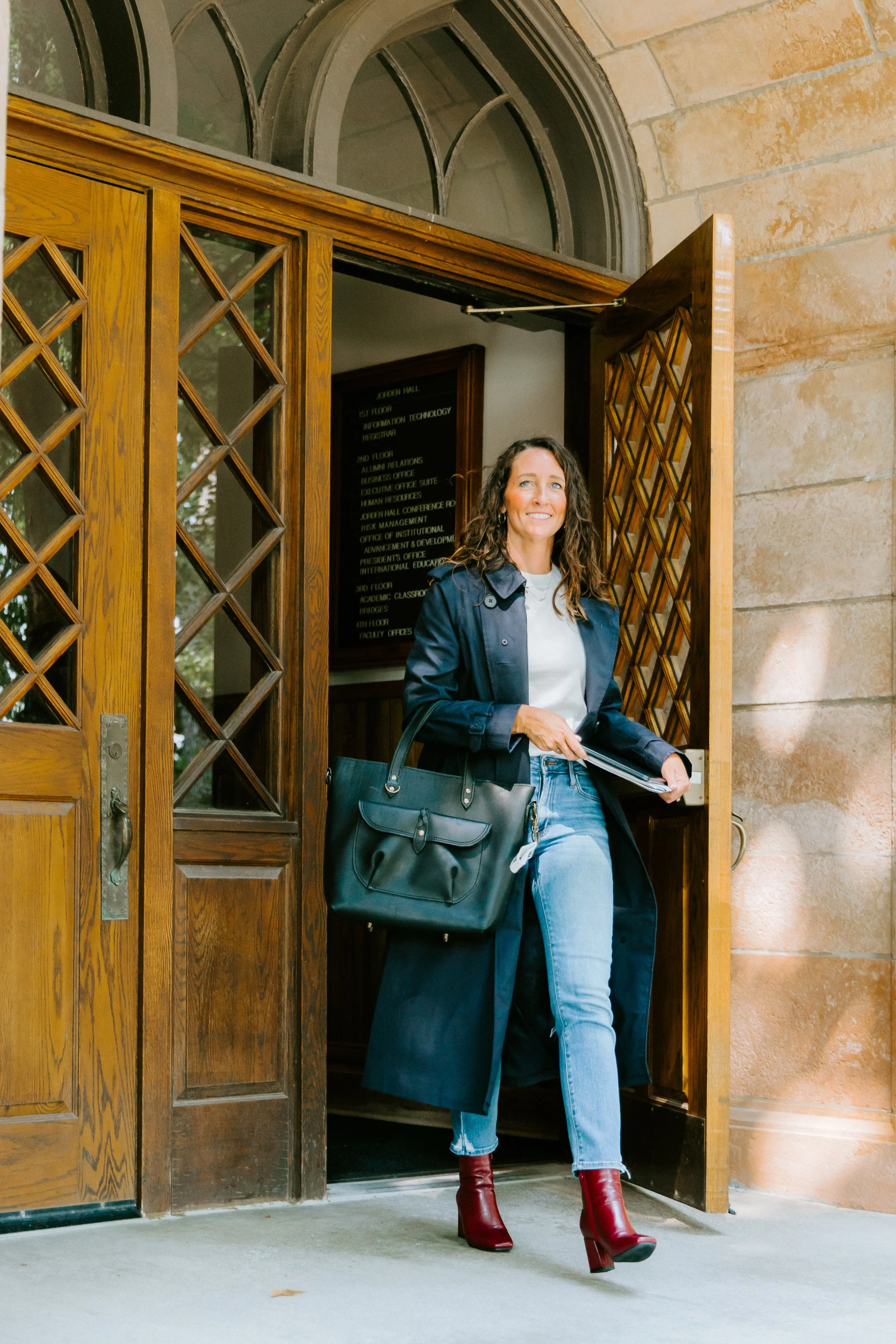 Woman exiting a building through a wooden door, wearing a navy coat, white shirt, blue jeans, and red high-heeled boots, holding a black handbag and some papers.