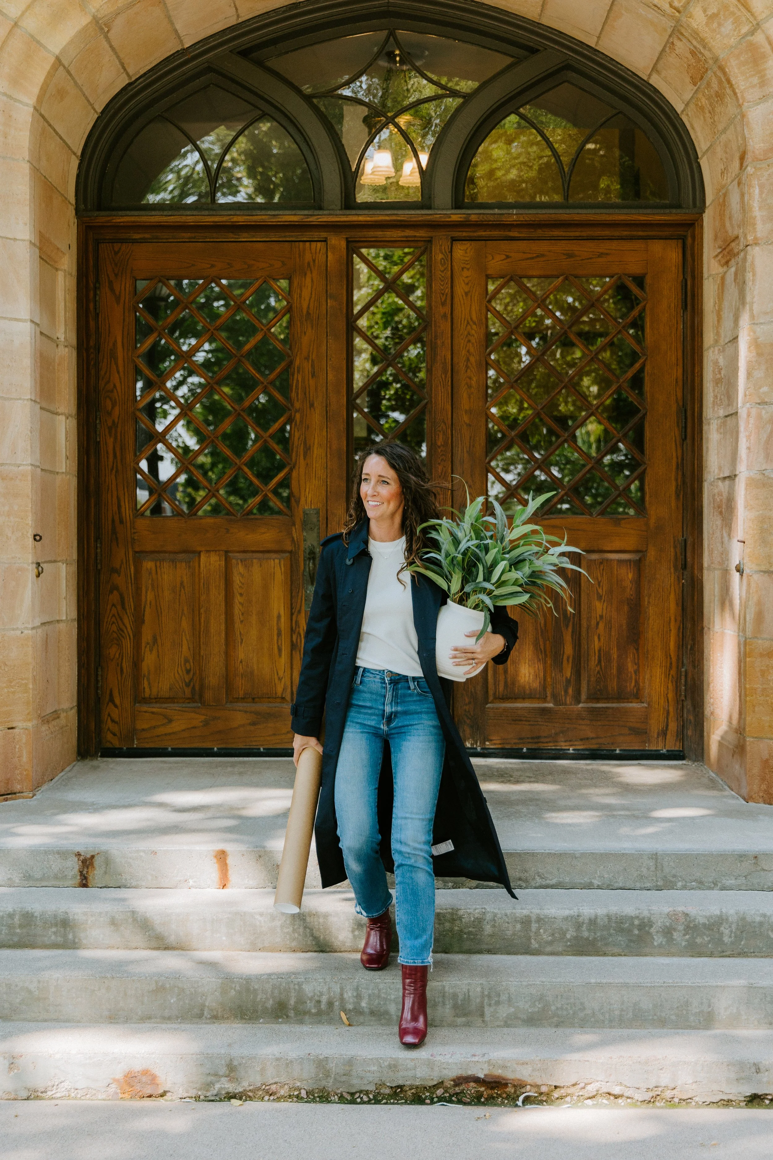Woman walking out of a building with large wooden double doors and a decorative arched window above, holding a potted plant in one hand and a rolled-up paper in the other, wearing a long black coat, jeans, red boots, and a white top.