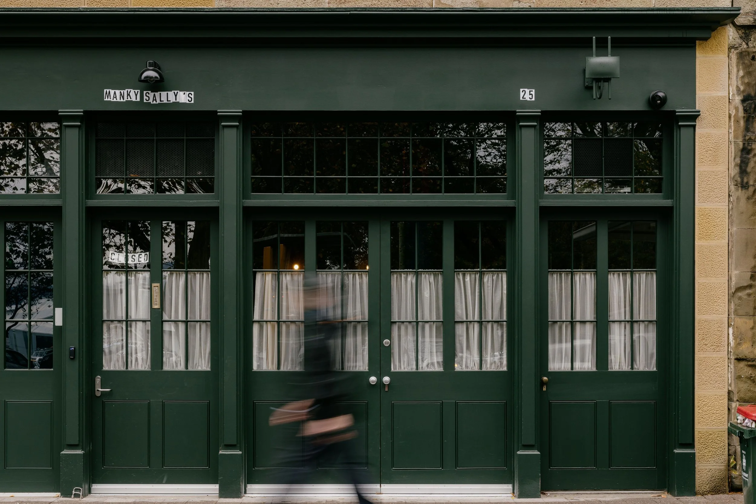 Green storefront with curtains behind glass doors, sign saying 'Manky Sally's', and a person walking by blurred due to motion.