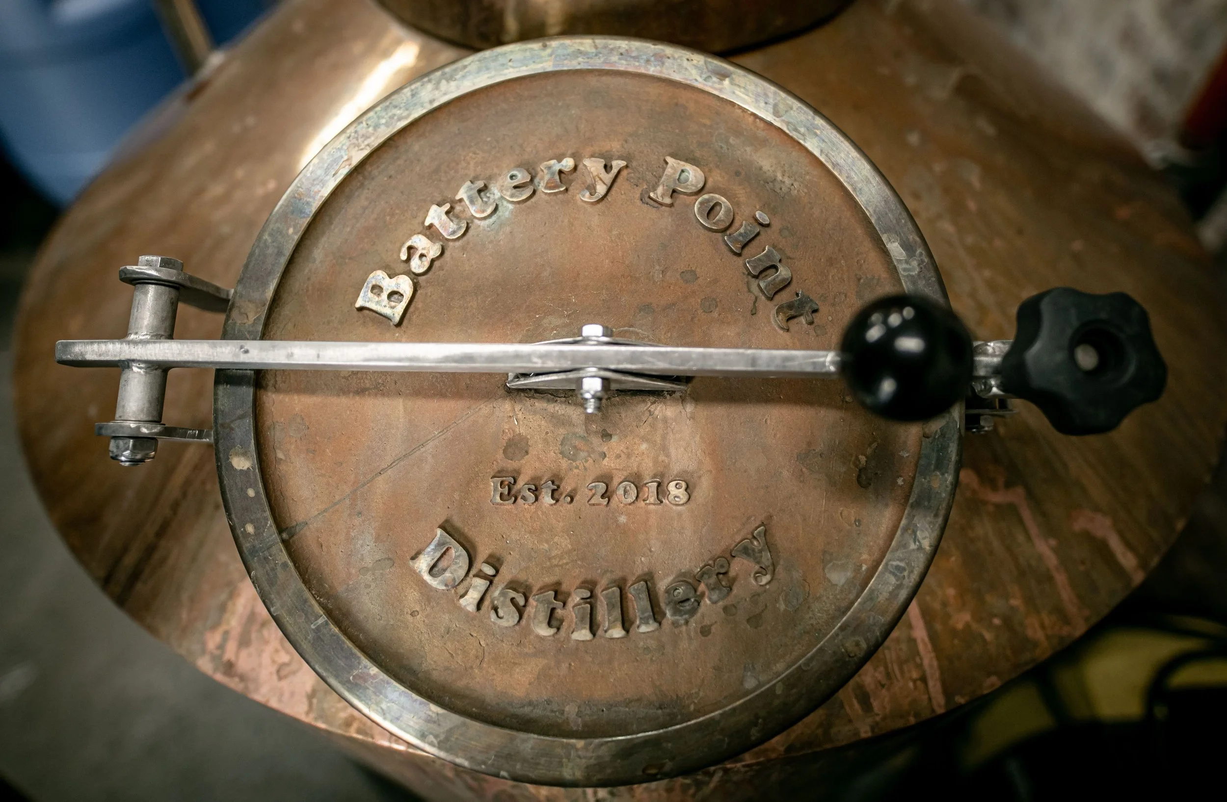 Top view of a vintage distillery copper drum with the words 'Batttery Point Distillers' and 'Est. 2018' engraved on it, showing a metal lid and a valve with a black knob.