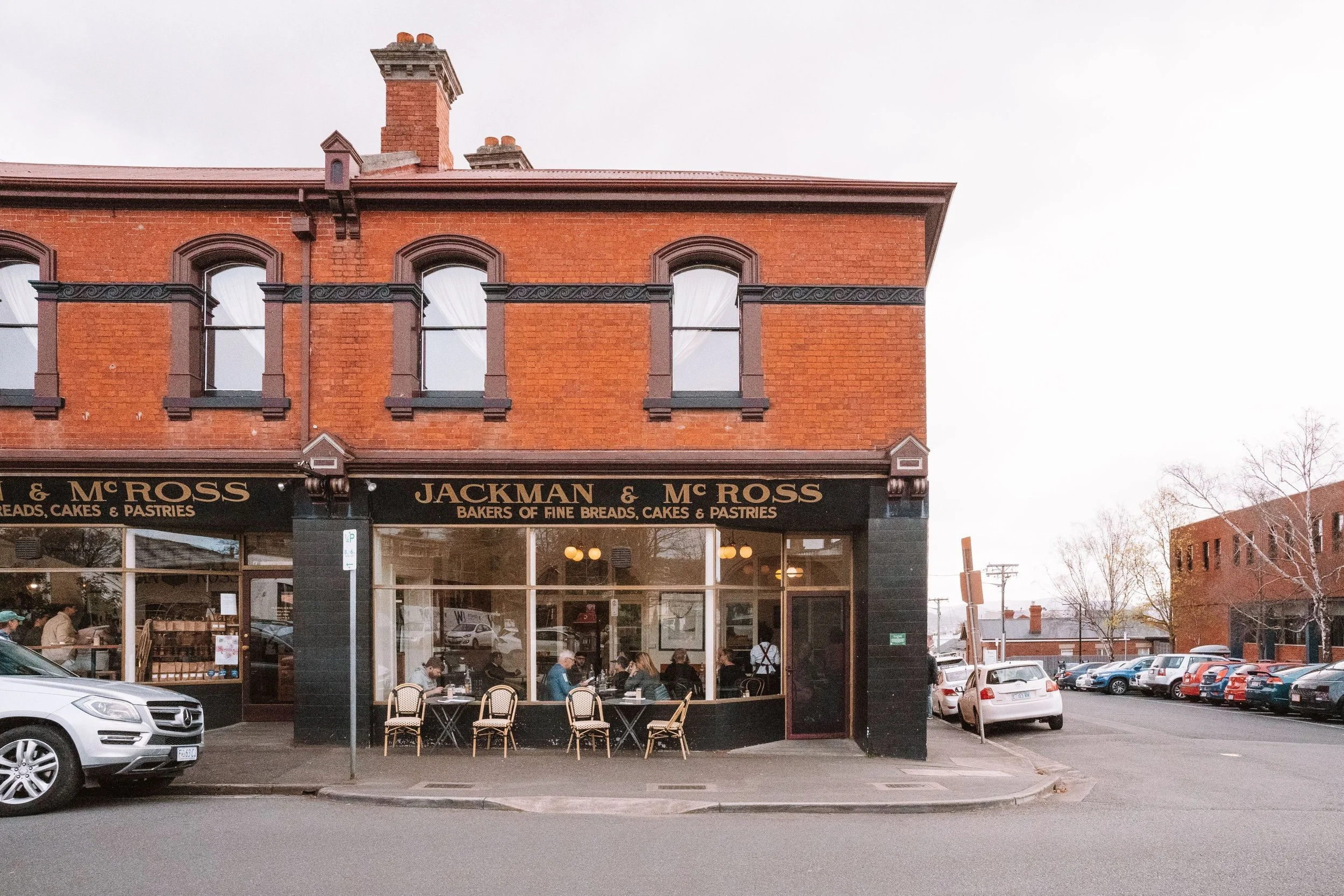 A two-story red brick bakery building with black trim, labeled 'Jackman & McRoss Bakers of Fine Breads, Cakes & Pastries,' with outdoor seating and parked cars in front.