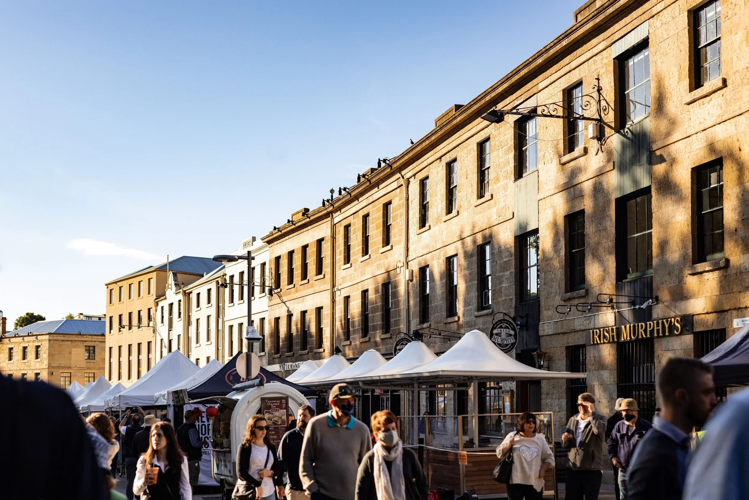 Street market scene with tents and a crowd of people walking in front of historic buildings with signs for Irish Murphy's, under a clear sky.