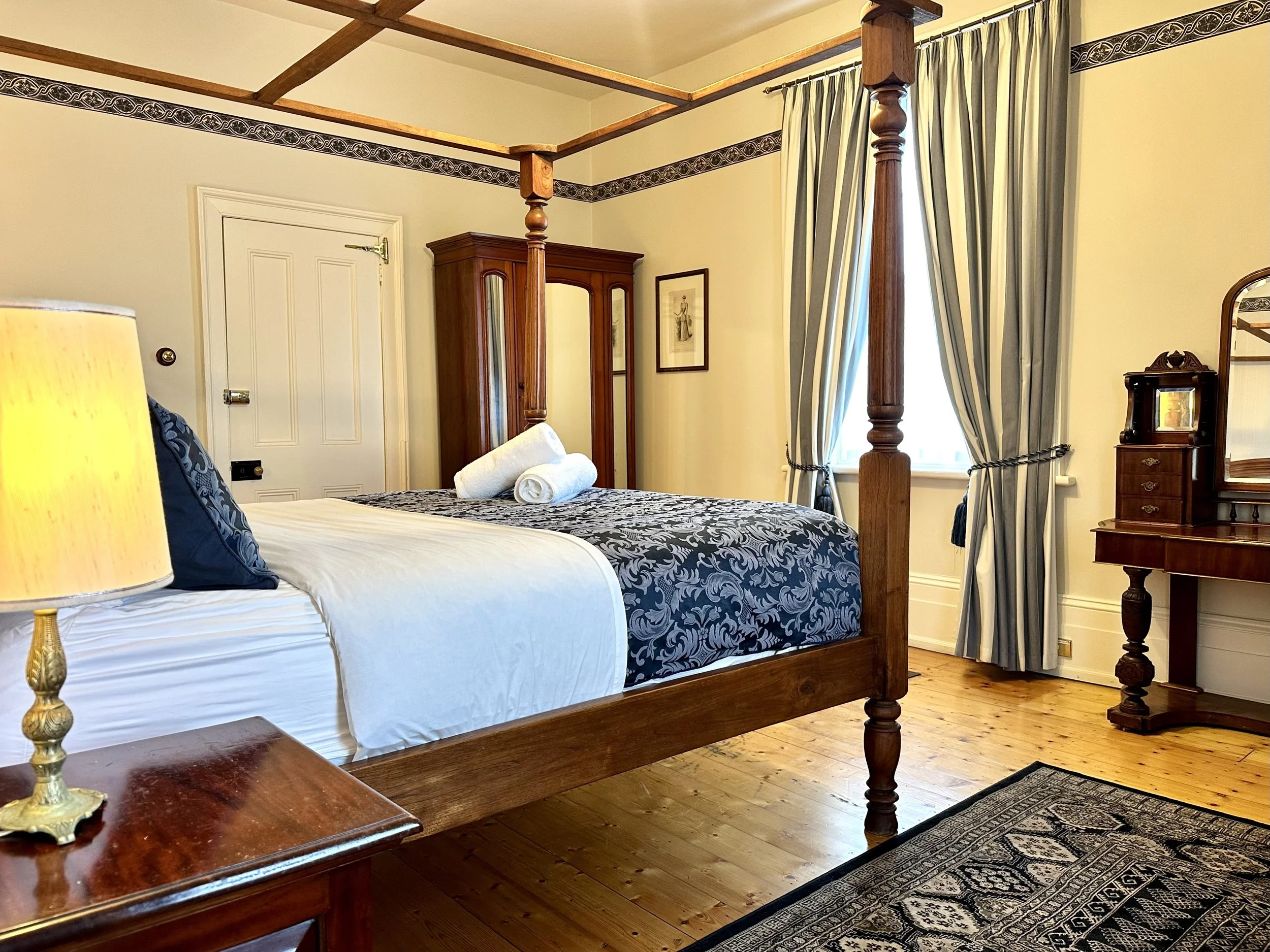 A bedroom with a four-poster wooden bed, featuring a white sheet and a floral dark blue bedspread, two rolled towels on top, a wooden dressing table with a mirror, a wooden wardrobe, a window with striped curtains, a side table with a lamp, and a pat