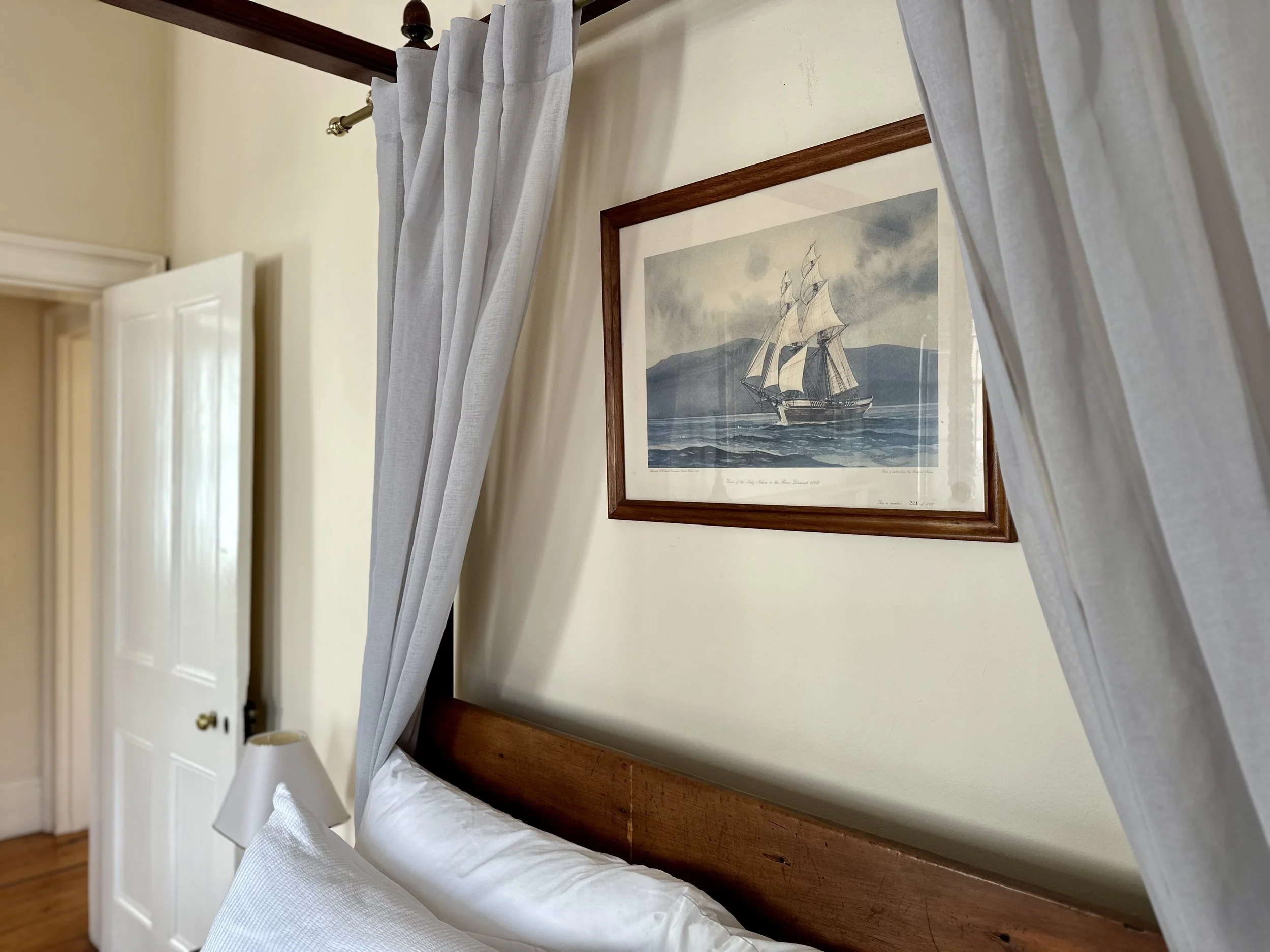 A bedroom with a wooden headboard, white bedding, white curtains, and a framed picture of a sailing ship on the wall.