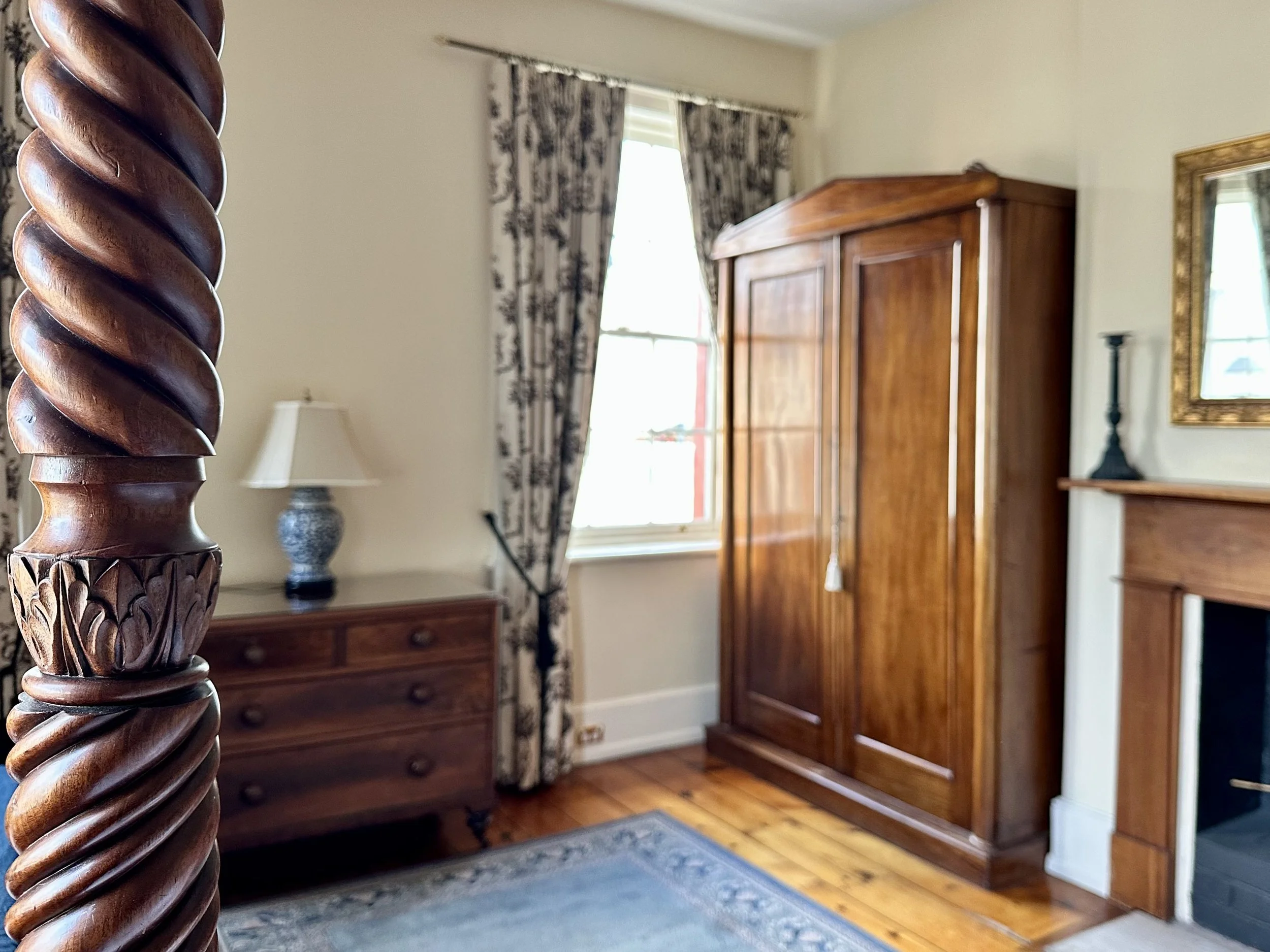 Part of a wooden four-poster bedpost in the foreground, with a bedroom featuring a wooden wardrobe, a dresser with a lamp, a window with floral curtains, and a mirror on the wall.