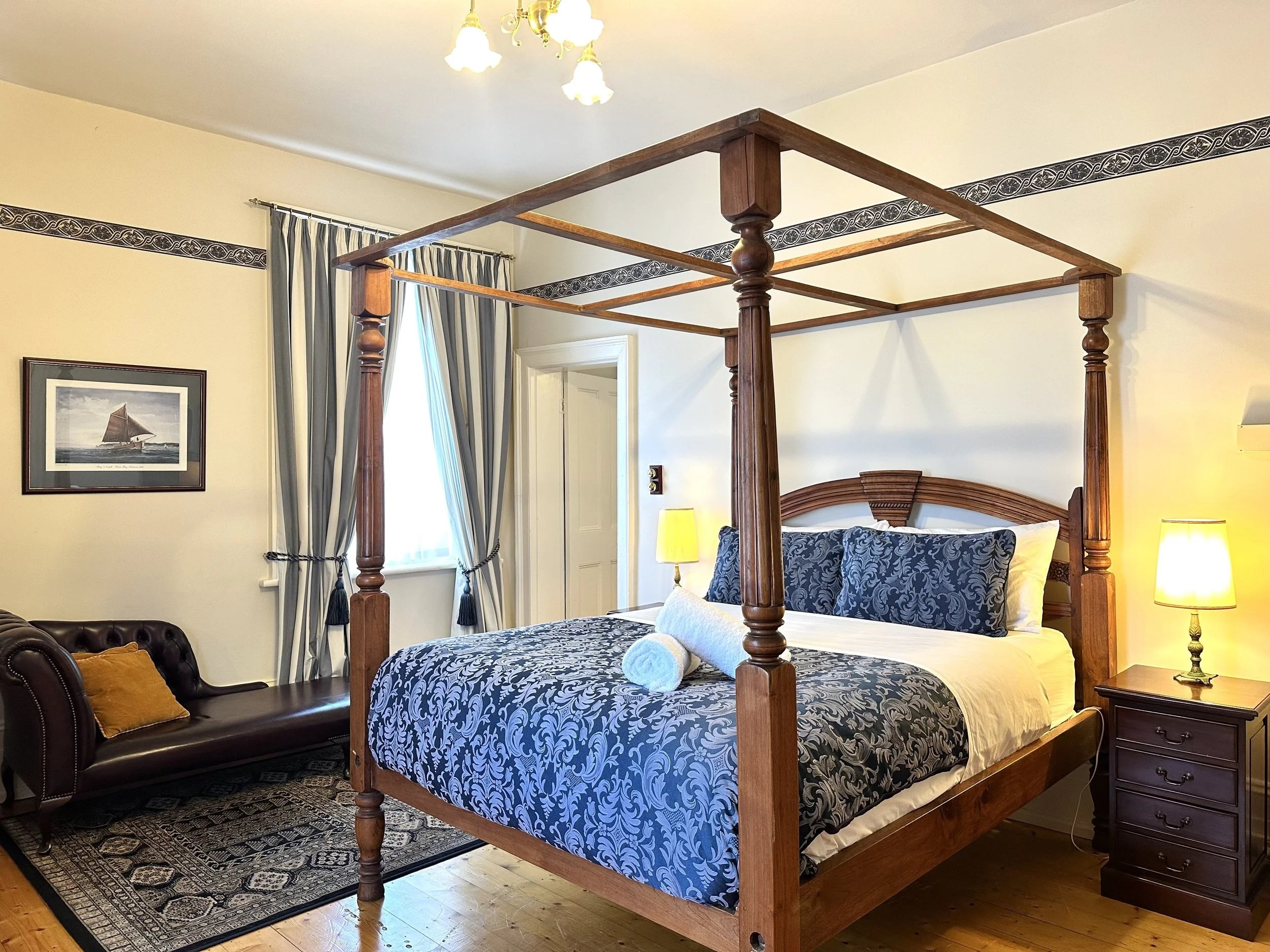 A bedroom with a wooden canopy bed, dark blue patterned bedding, and matching pillows. There are two yellow table lamps on dark wood nightstands, a leather tufted bench, and striped curtains near a window. A framed picture of a sailboat hangs on the