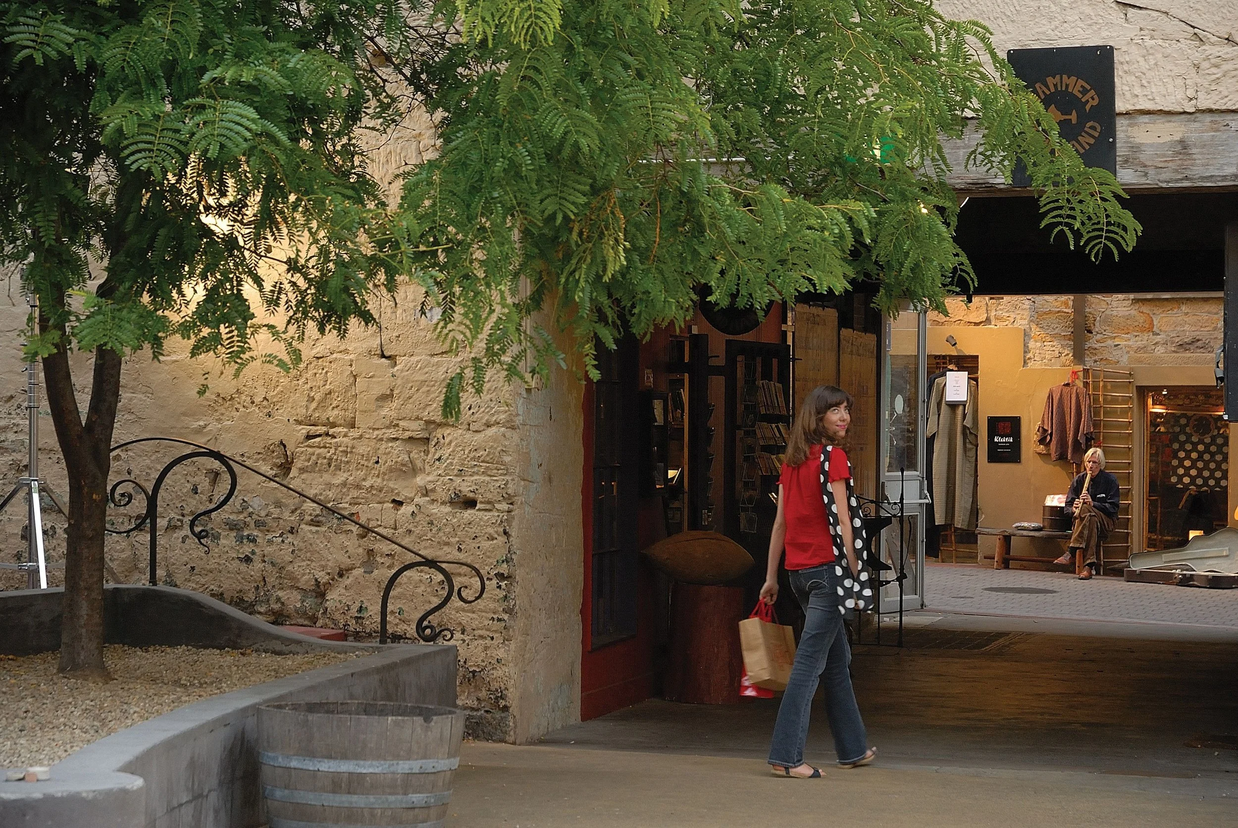 A woman with shoulder-length brown hair, wearing a red shirt, jeans, and sandals, is walking past an entrance to a shop or gallery. She is carrying shopping bags. The background includes a man sitting inside playing a wind instrument, and a wooden an