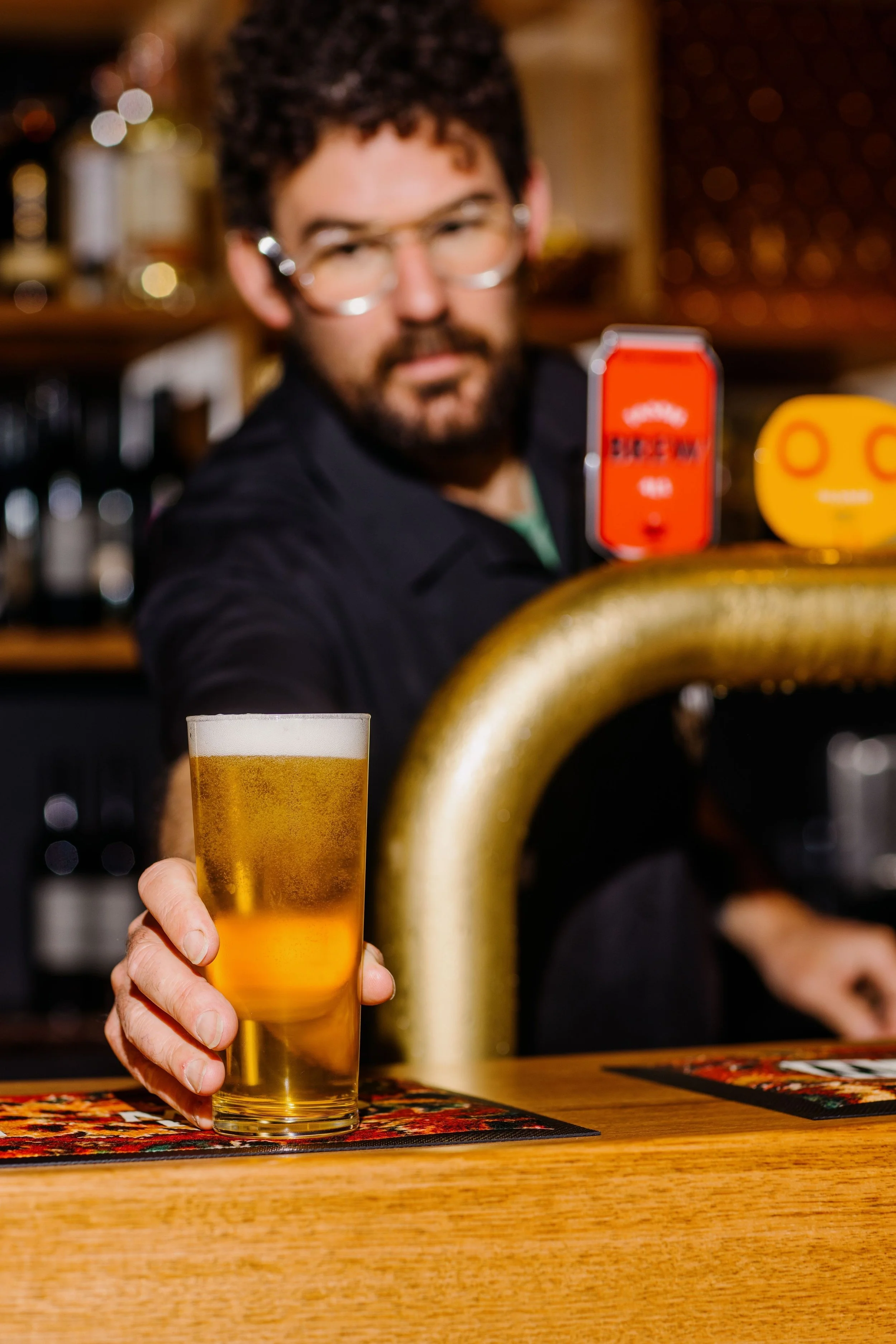 A bartender presenting a glass of beer at a bar with a golden tap, a red soda machine, and a yellow device on the counter.