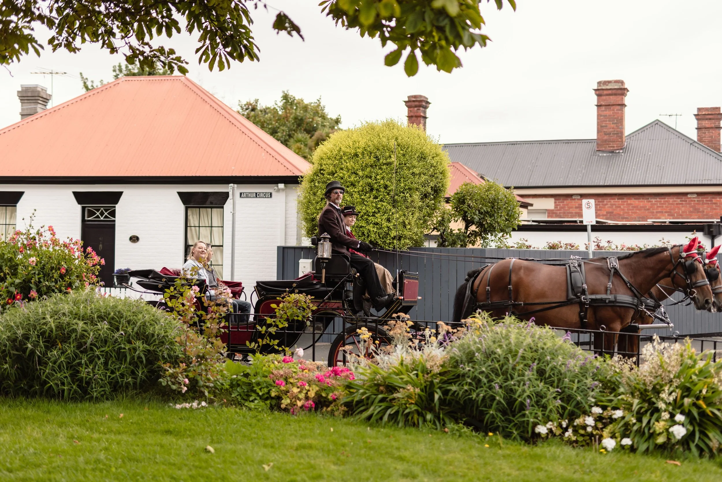A horse-drawn carriage with two people in vintage clothing, a man and a woman, riding through a garden with colorful flowers. In the background, there are houses with red and gray roofs and brick chimneys.