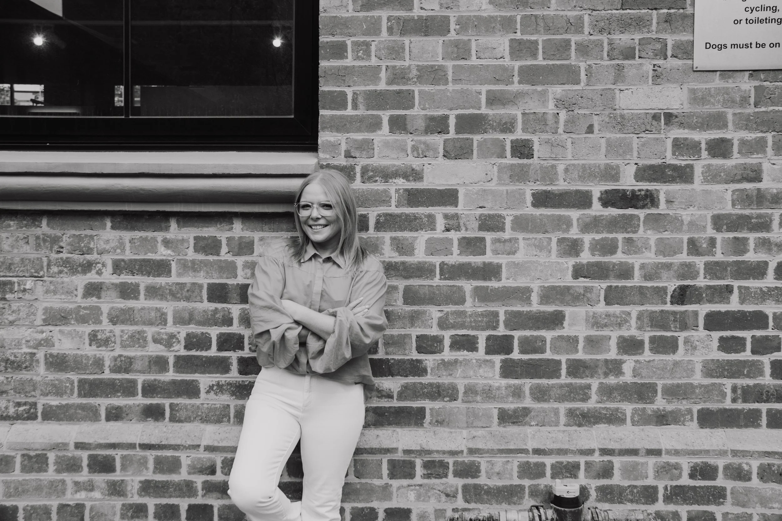 A woman with glasses, wearing a light-colored shirt and pants, standing with her arms crossed and smiling in front of a brick wall.