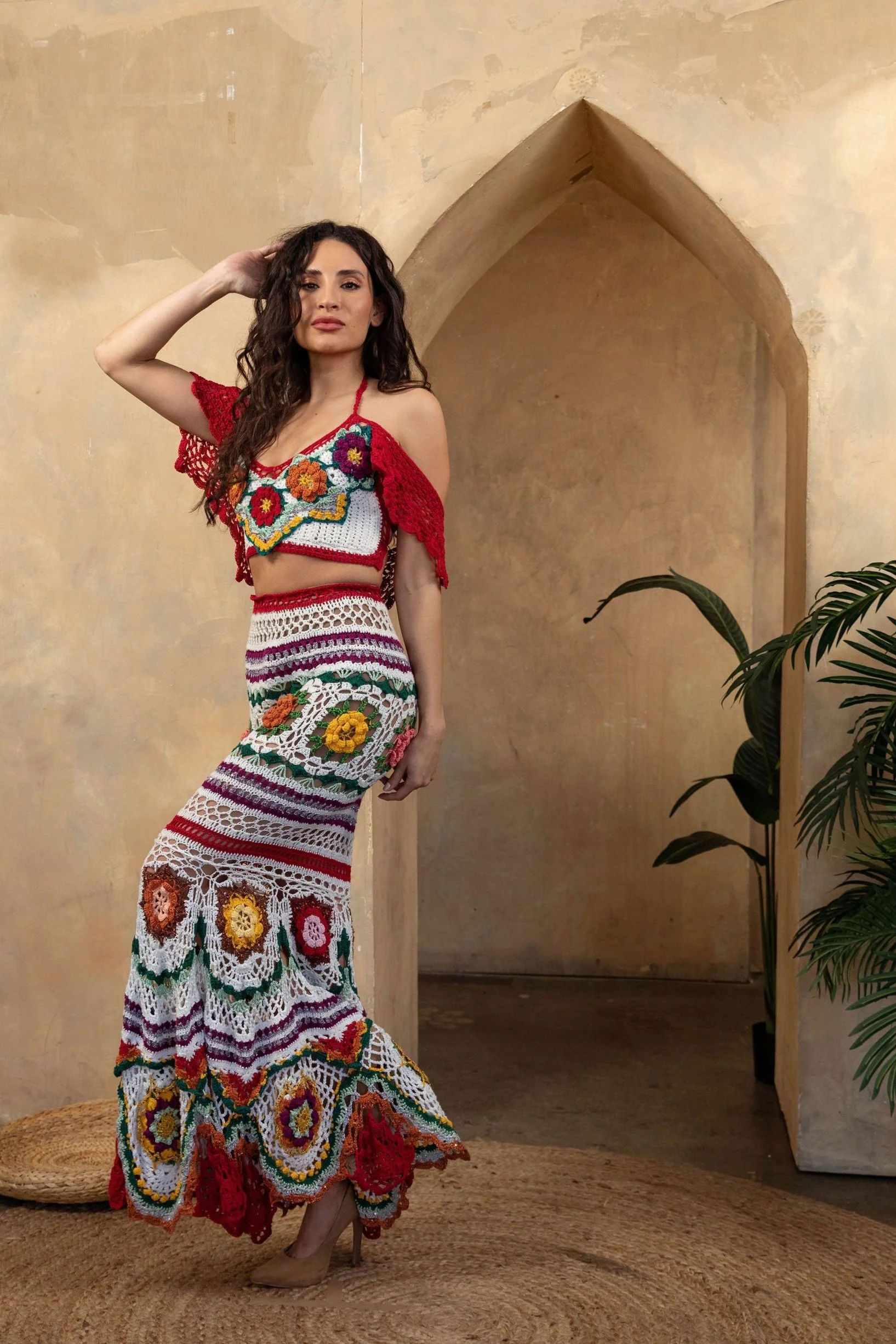 A woman wearing a colorful, crocheted bohemian-style outfit consisting of a crop top and a long skirt with floral patterns, standing in front of a textured beige wall with an arched niche and a plant nearby.