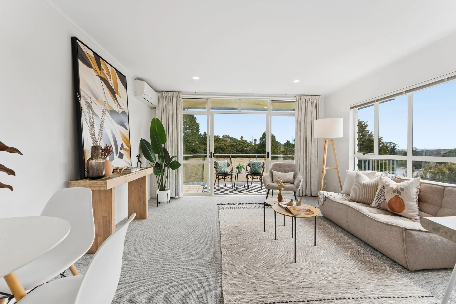 Living room with beige carpet, wooden dining table, and chairs, beige sofas, large windows with outdoor greenery, and a striped rug.
