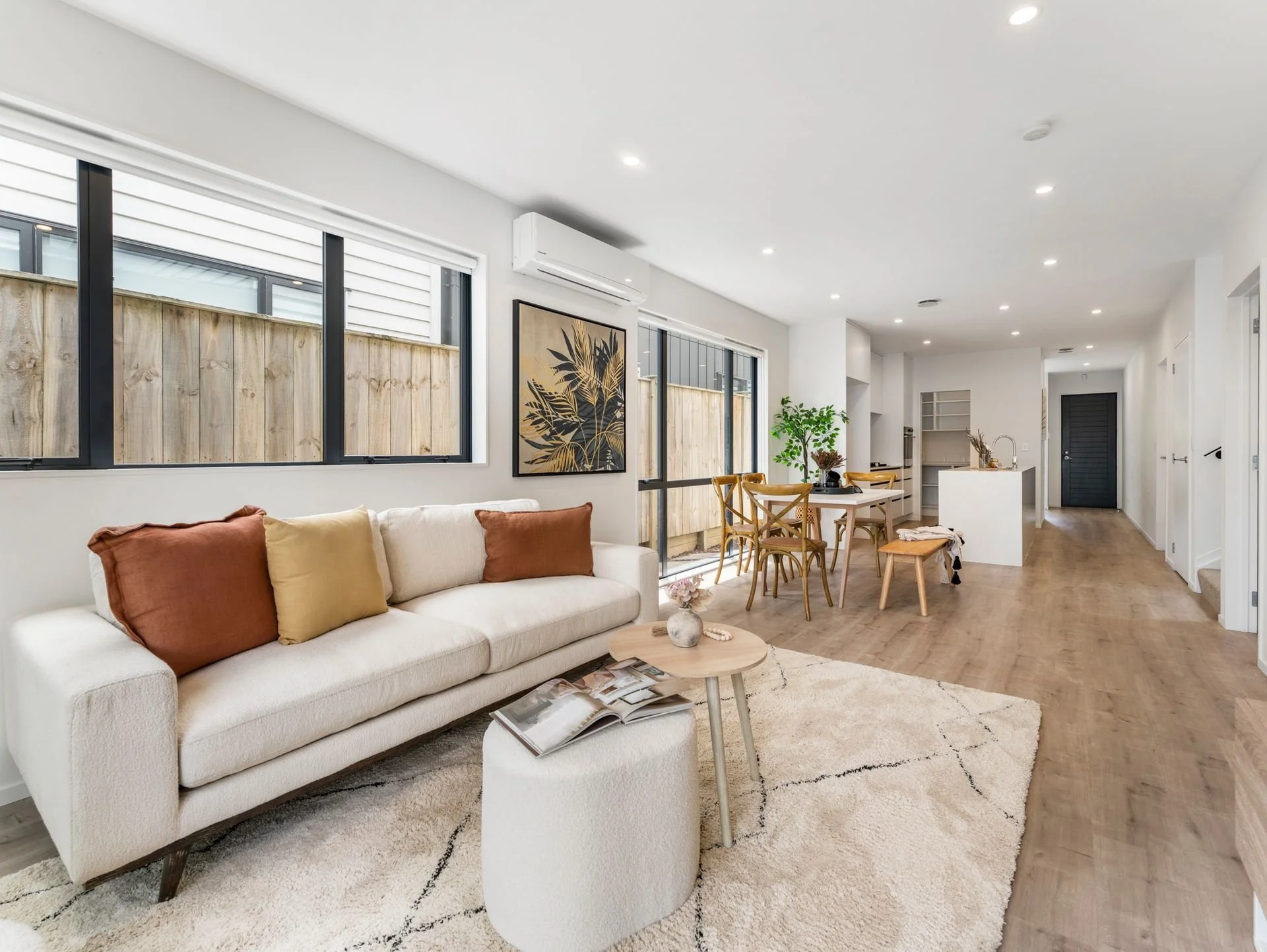 Living room with white sofa, colorful throw pillows, wooden coffee table with magazines, beige rug, multiple windows, art on wall, and open kitchen and dining area with wooden table and chairs