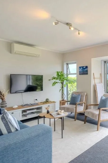 Living room with a wall-mounted TV, a beige armchair, a blue sofa, a small round coffee table, a potted plant, windows letting in natural light, and a sliding glass door leading outside.