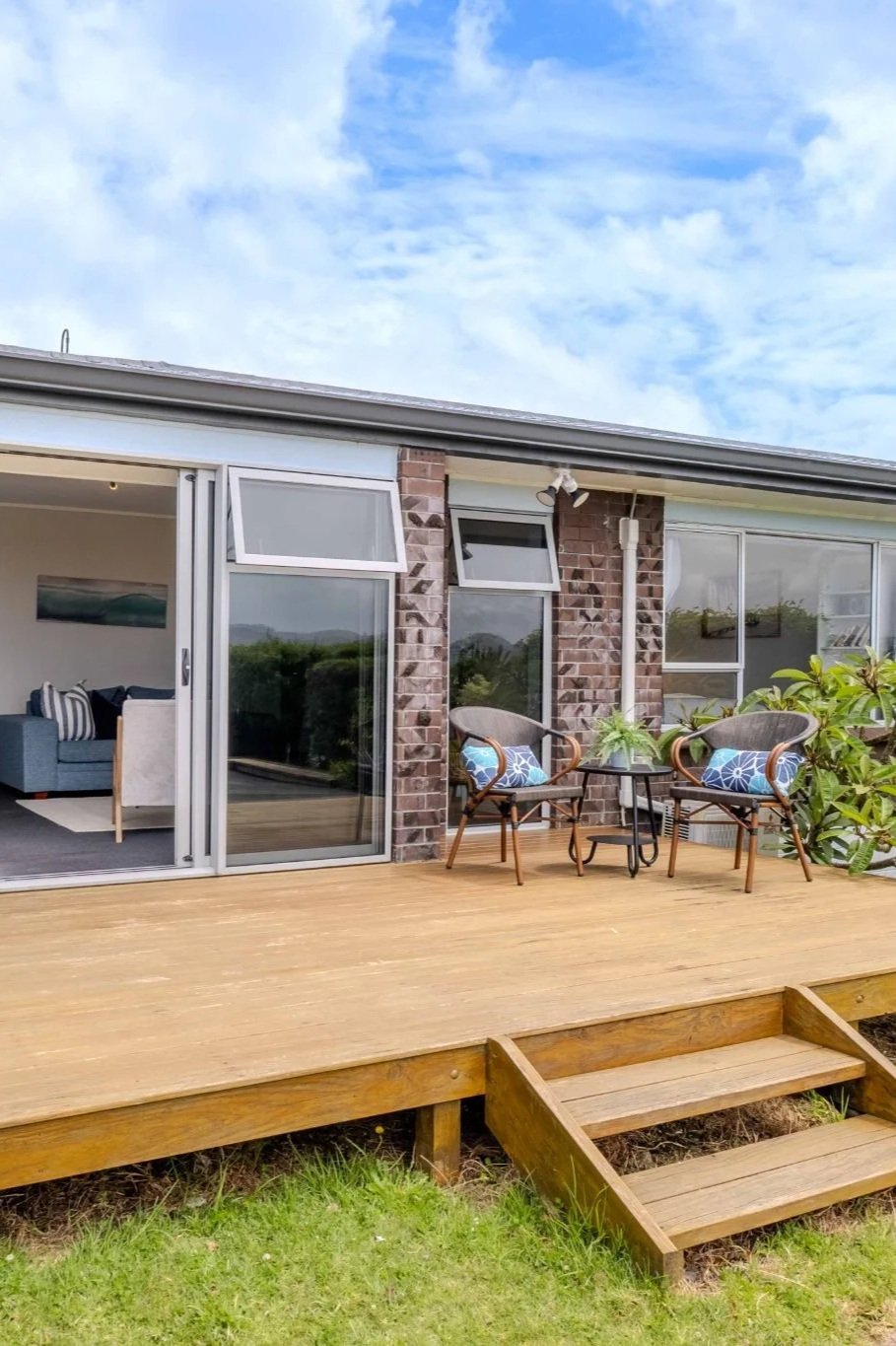 Outdoor wooden deck with two chairs and a small table, attached to a house with brick and glass sliding door, overlooking a garden and cloudy sky.