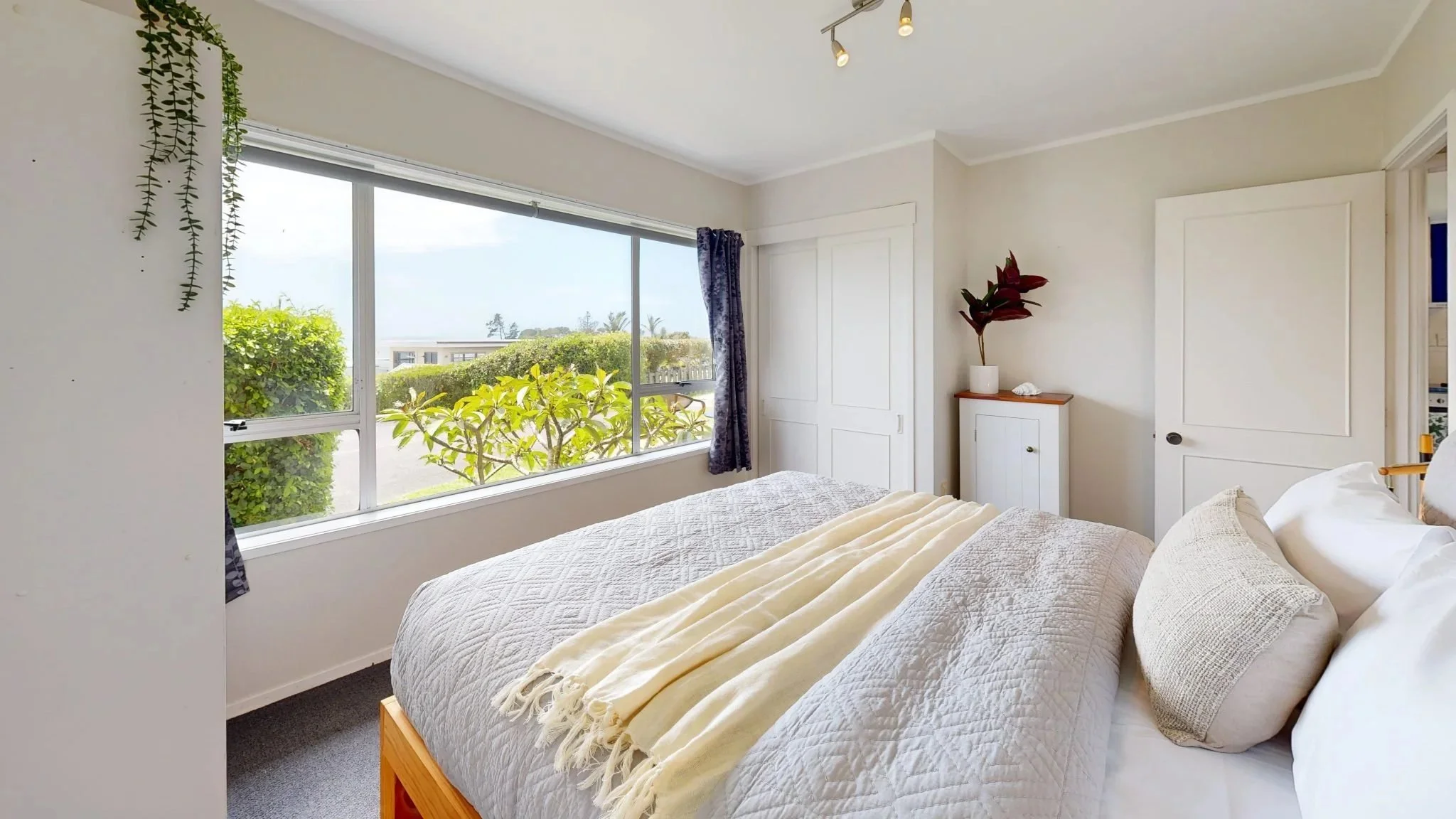 A bedroom with a large window showing outside greenery, a white quilted bed with pillows and a cream blanket, a small white cabinet with a potted plant, and a door leading to a closet or another room.