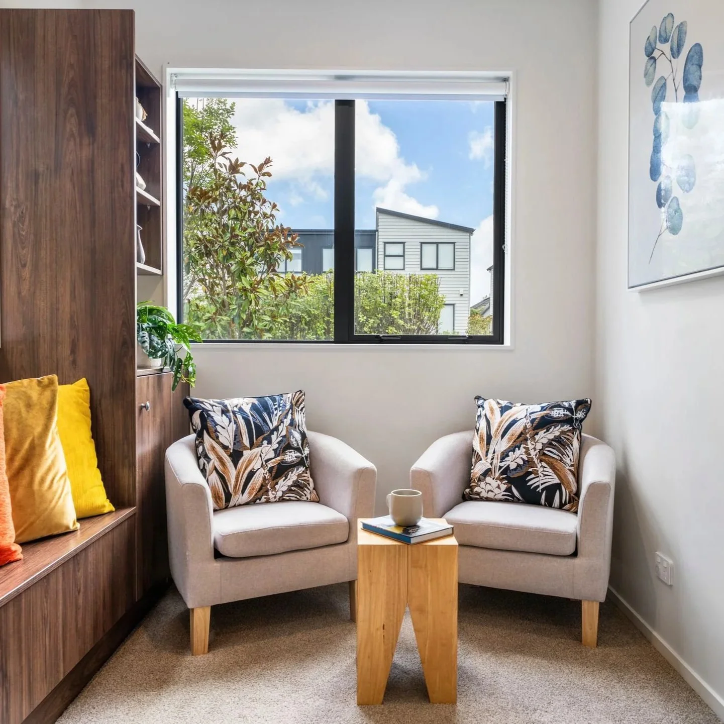 A cozy sitting area with two white armchairs, each with patterned pillows, a small wooden side table with a mug and books, against a window with a view of trees and houses outside.