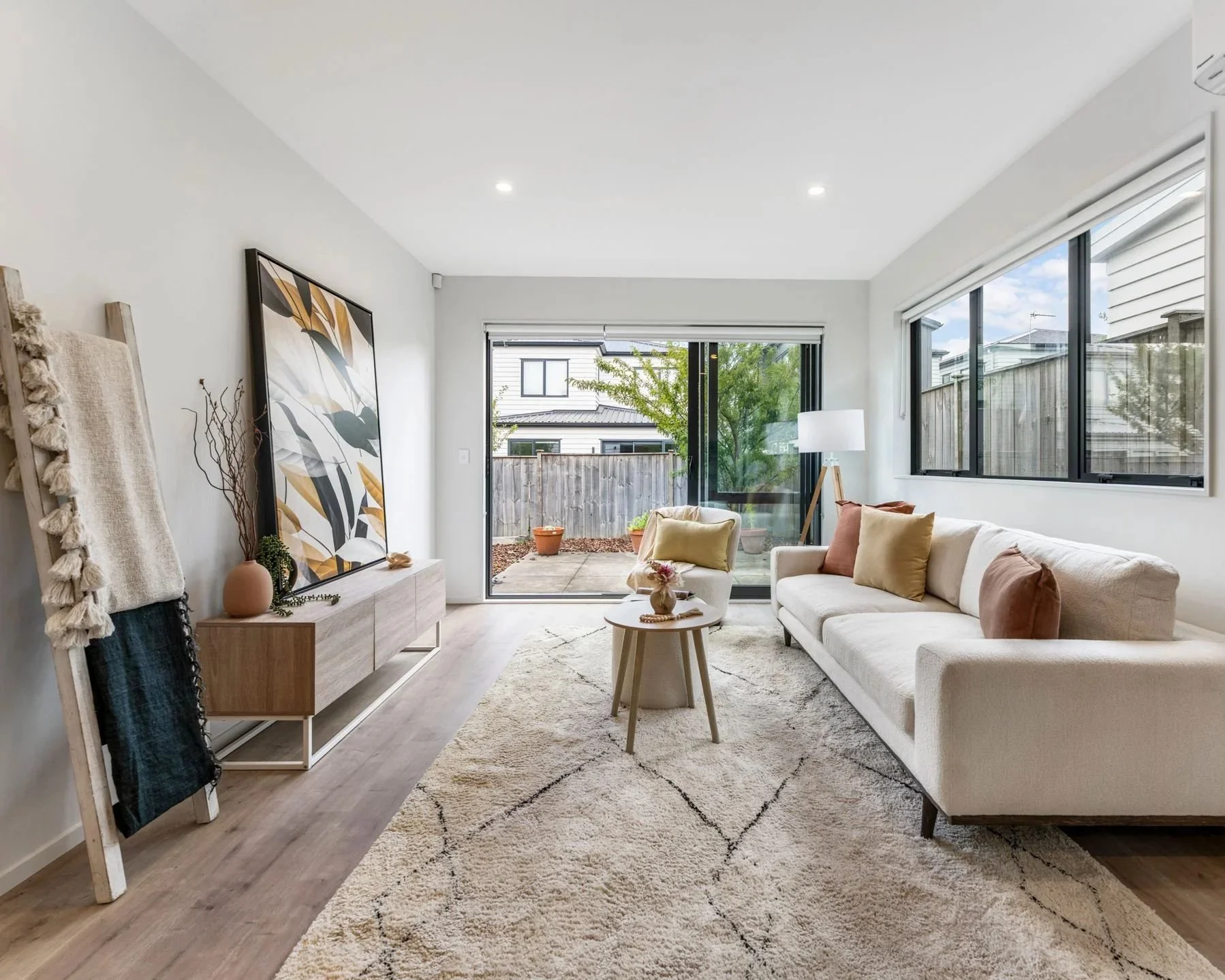 Bright living room with white walls, a cream sofa with pillows, a small round coffee table, large windows, and sliding glass door leading outside. There is a patterned beige rug, a floor lamp, and a wooden TV stand with a large framed art piece and d