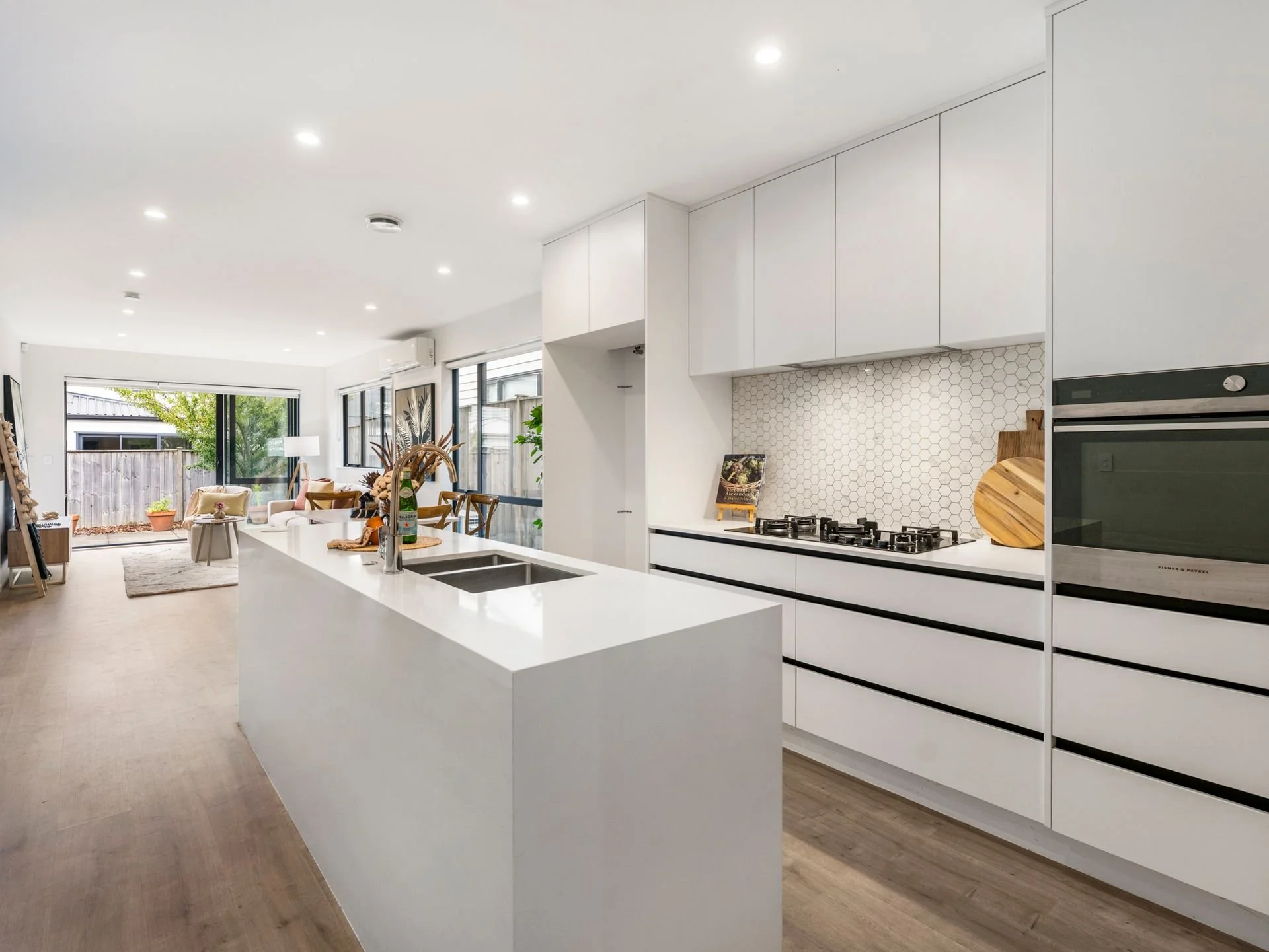 Modern open-plan kitchen with white cabinetry and island, hexagonal backsplash, built-in oven, and a living area with large windows and outdoor view.