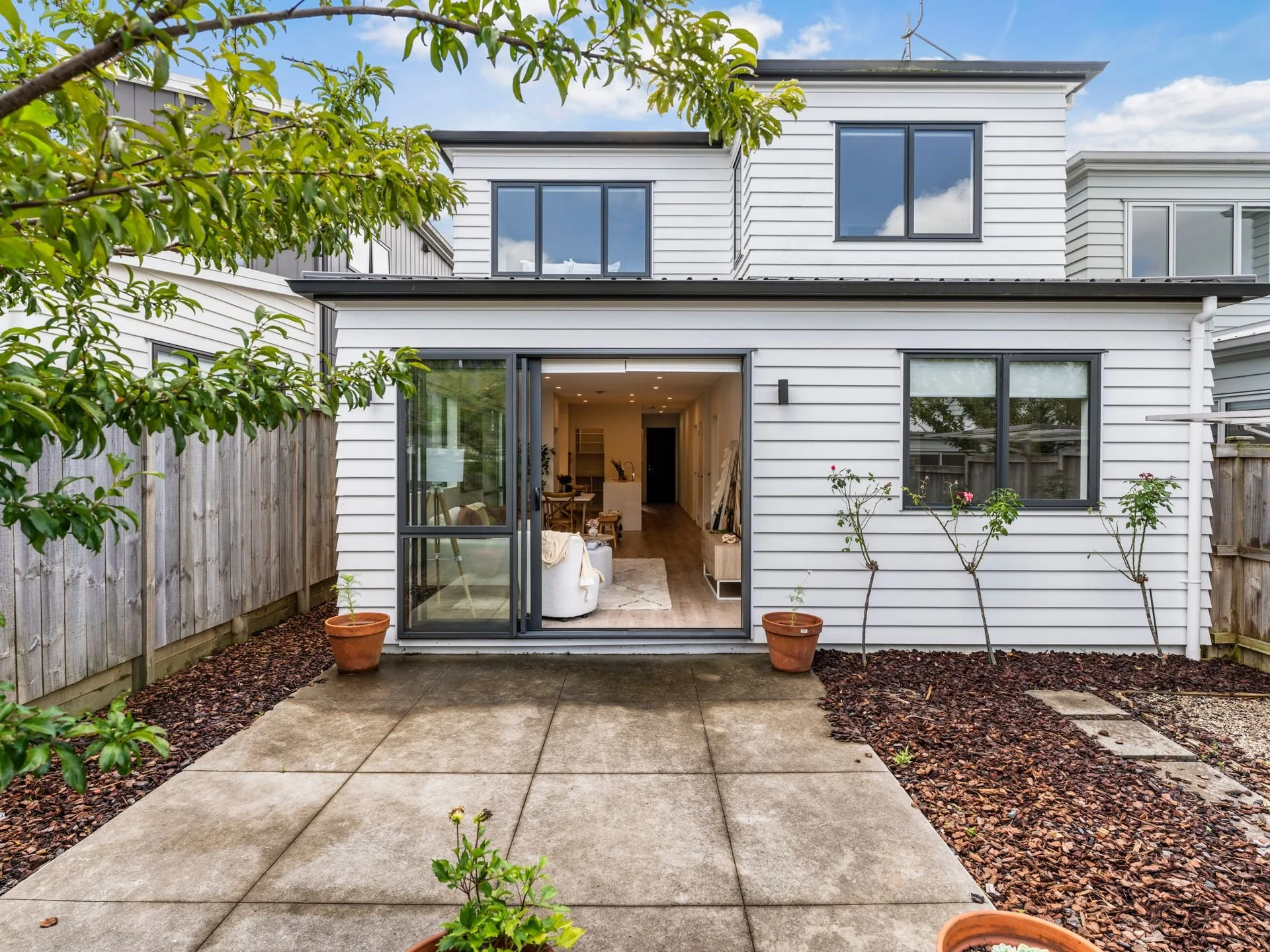 Backyard patio area with sliding glass door leading to interior living space, surrounded by potted plants, a young tree, and a wooden fence, with a white multi-story house in the background.